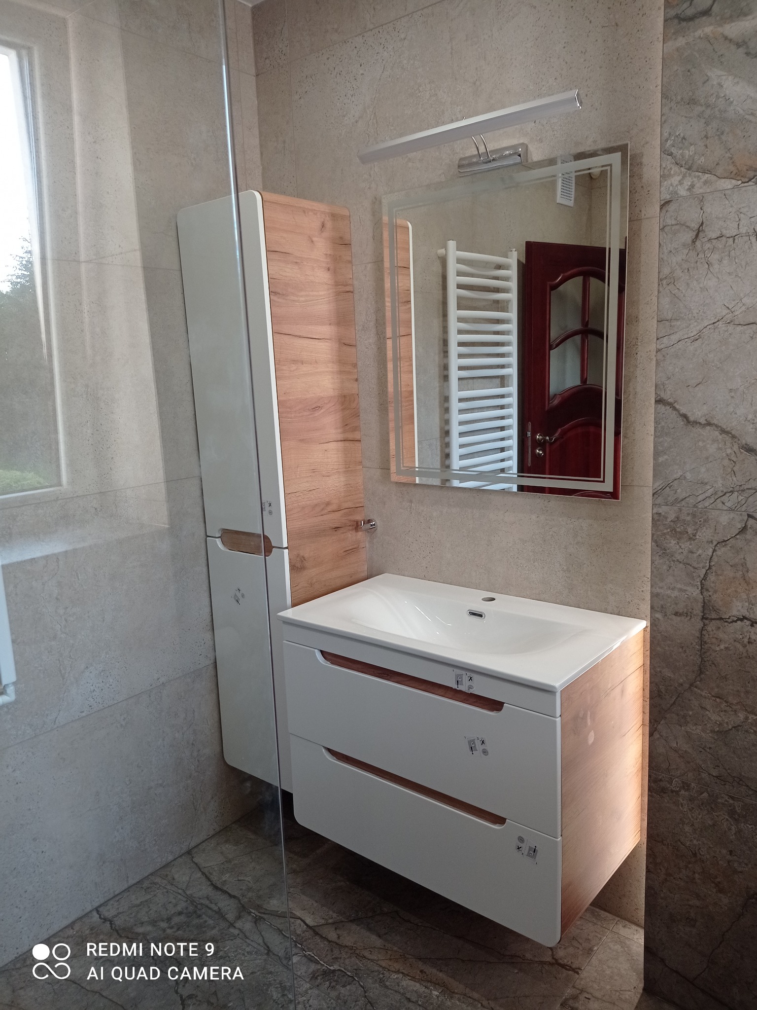 Nowoczesna łazienka z umywalką, szafką w odcieniach bieli i drewna. W lustrze odbija się grzejnik. Beżowe płytki na ścianach i podłodze.