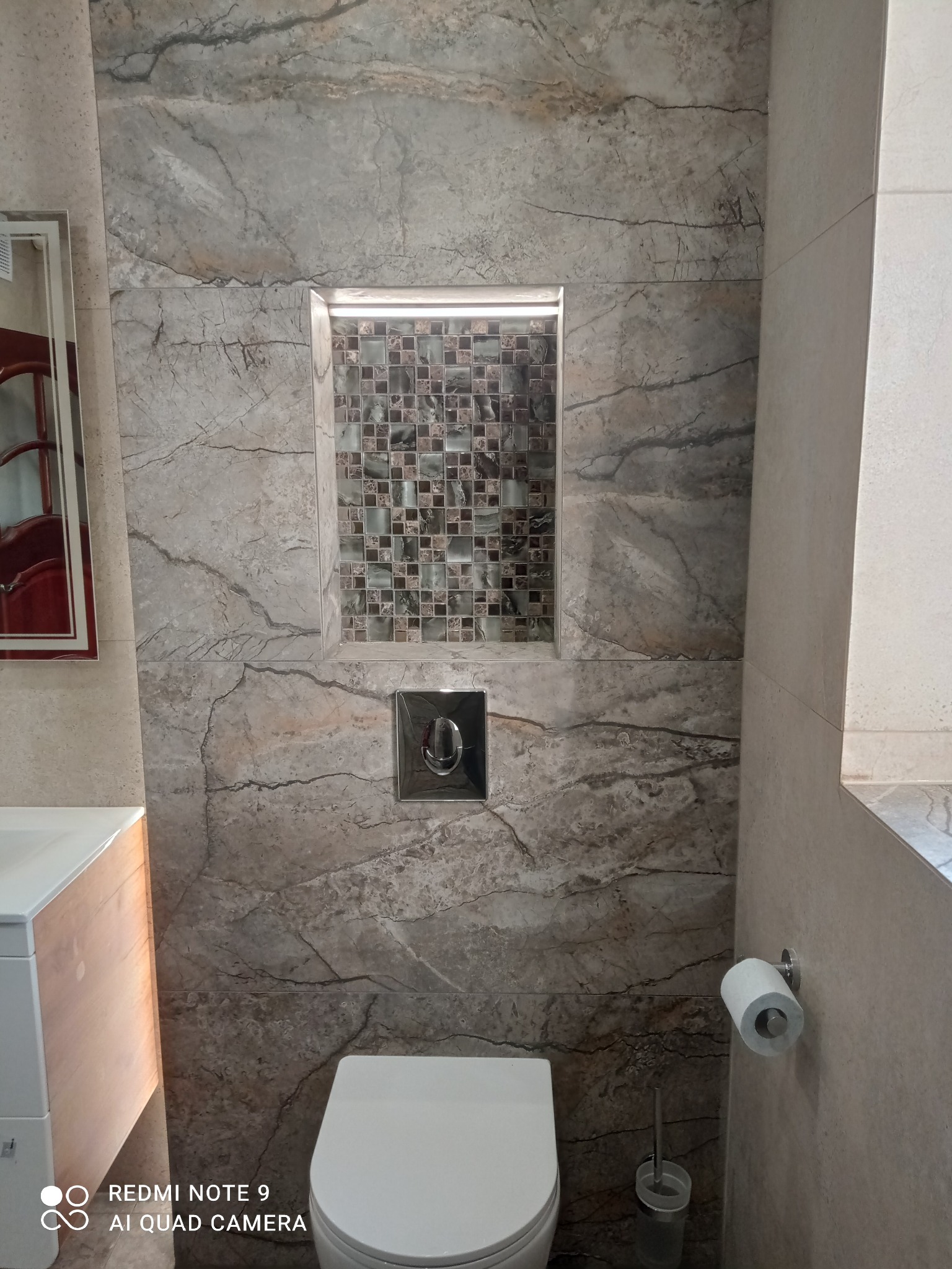 Wykończona łazienka z marmurowymi płytkami, wnęką z mozaiką i podświetleniem LED. Widoczna toaleta i fragment umywalki. Nowoczesny design z elementami dekoracyjnymi.