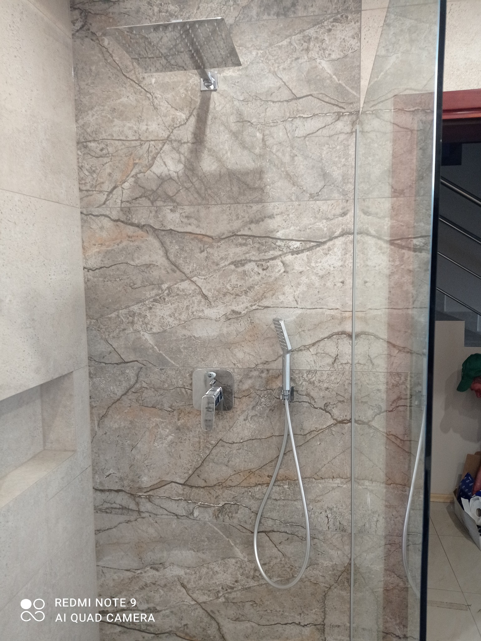 Kabina prysznicowa z deszczownicą i marmurową okładziną. Bateria podtynkowa, szklana ściana kabiny. Widok na schody w tle.