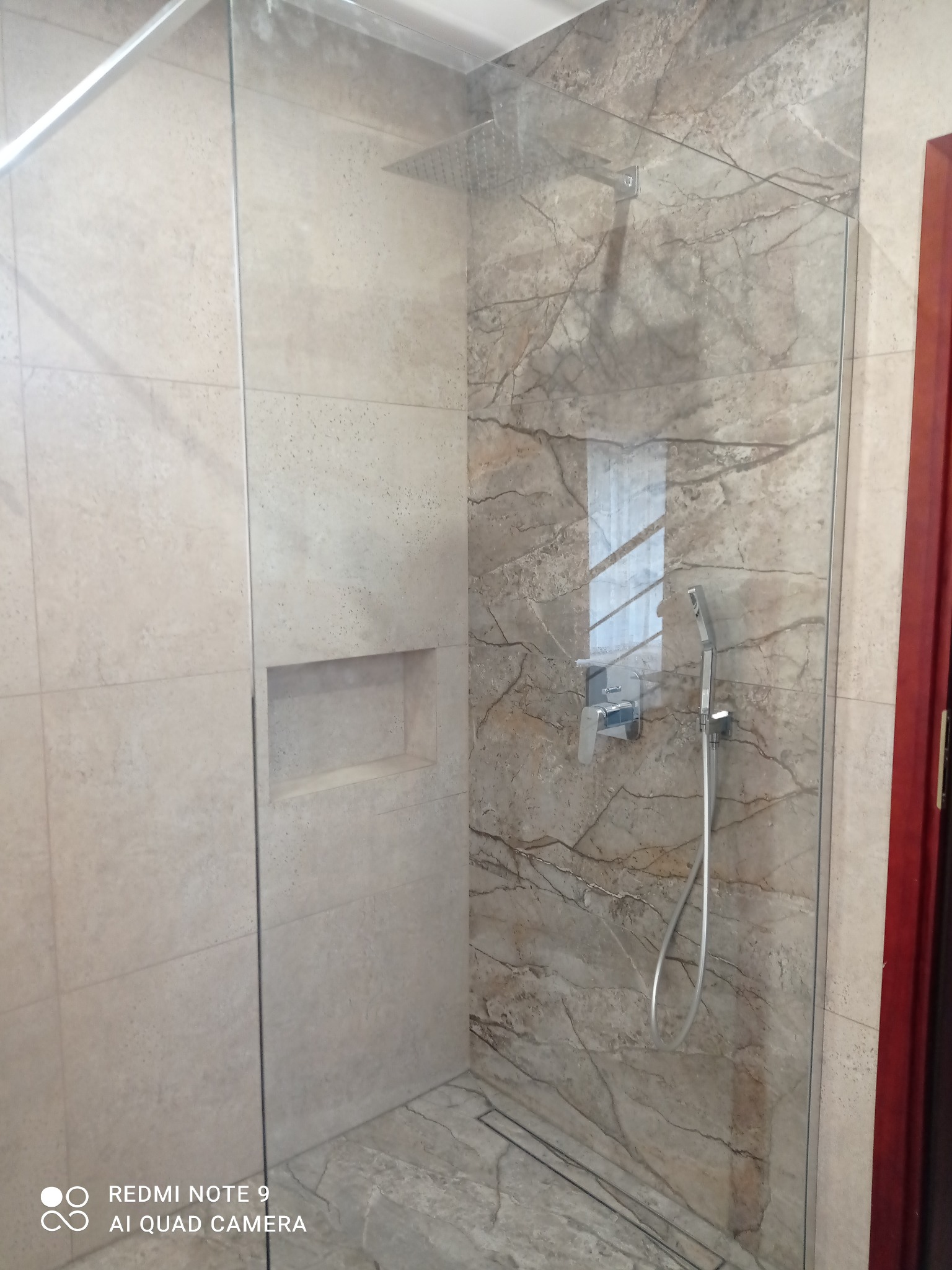 Kabina prysznicowa walk-in z marmurowymi płytkami, deszczownicą i wnęką w ścianie. Szklana tafla oddziela strefę mokrą od reszty łazienki. Widoczne detale baterii i wykończenia.