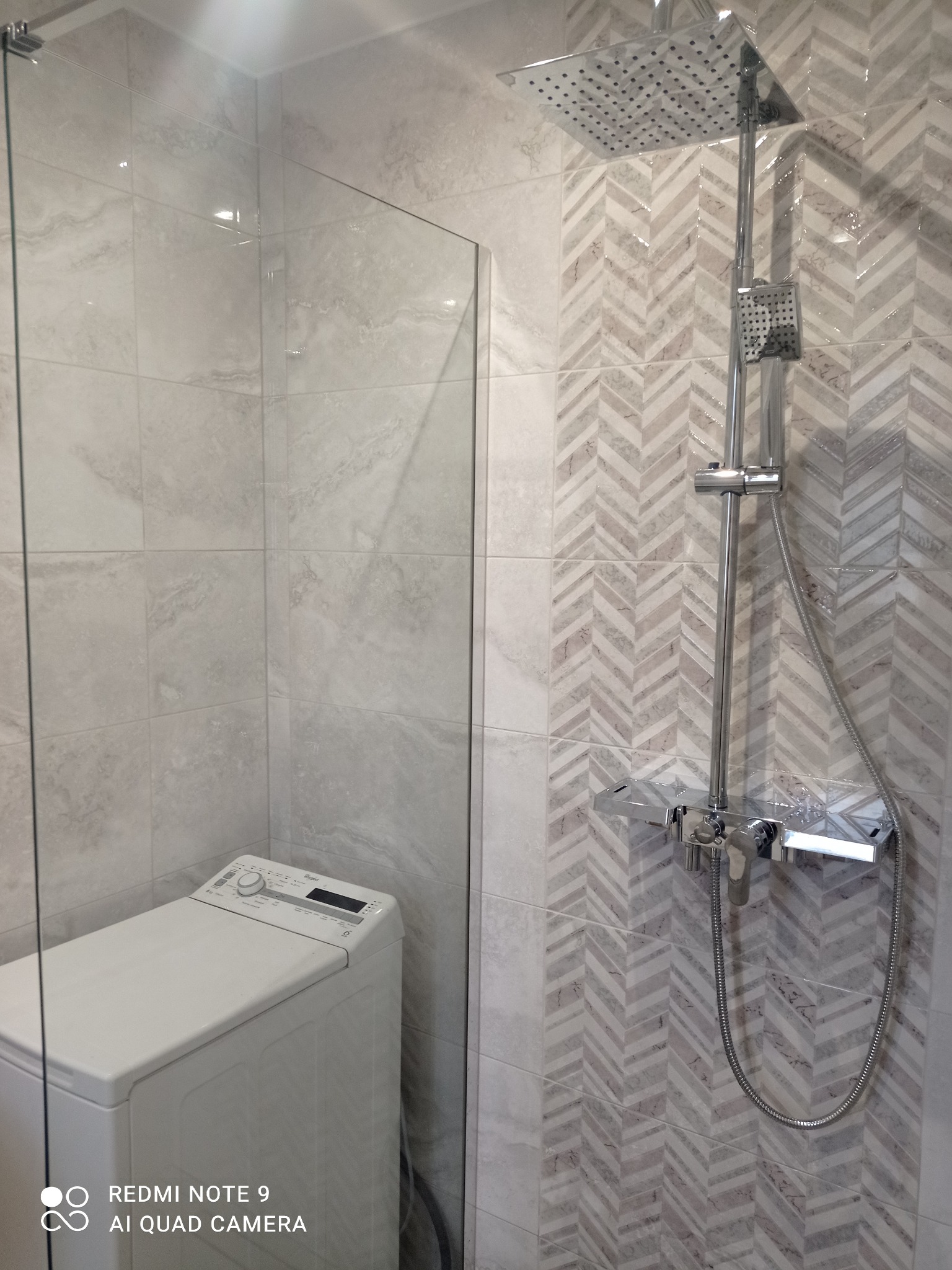 Nowoczesna łazienka z kabiną prysznicową typu walk-in, białą pralką i dekoracyjnymi płytkami w jodełkę na ścianie z chromowaną armaturą.