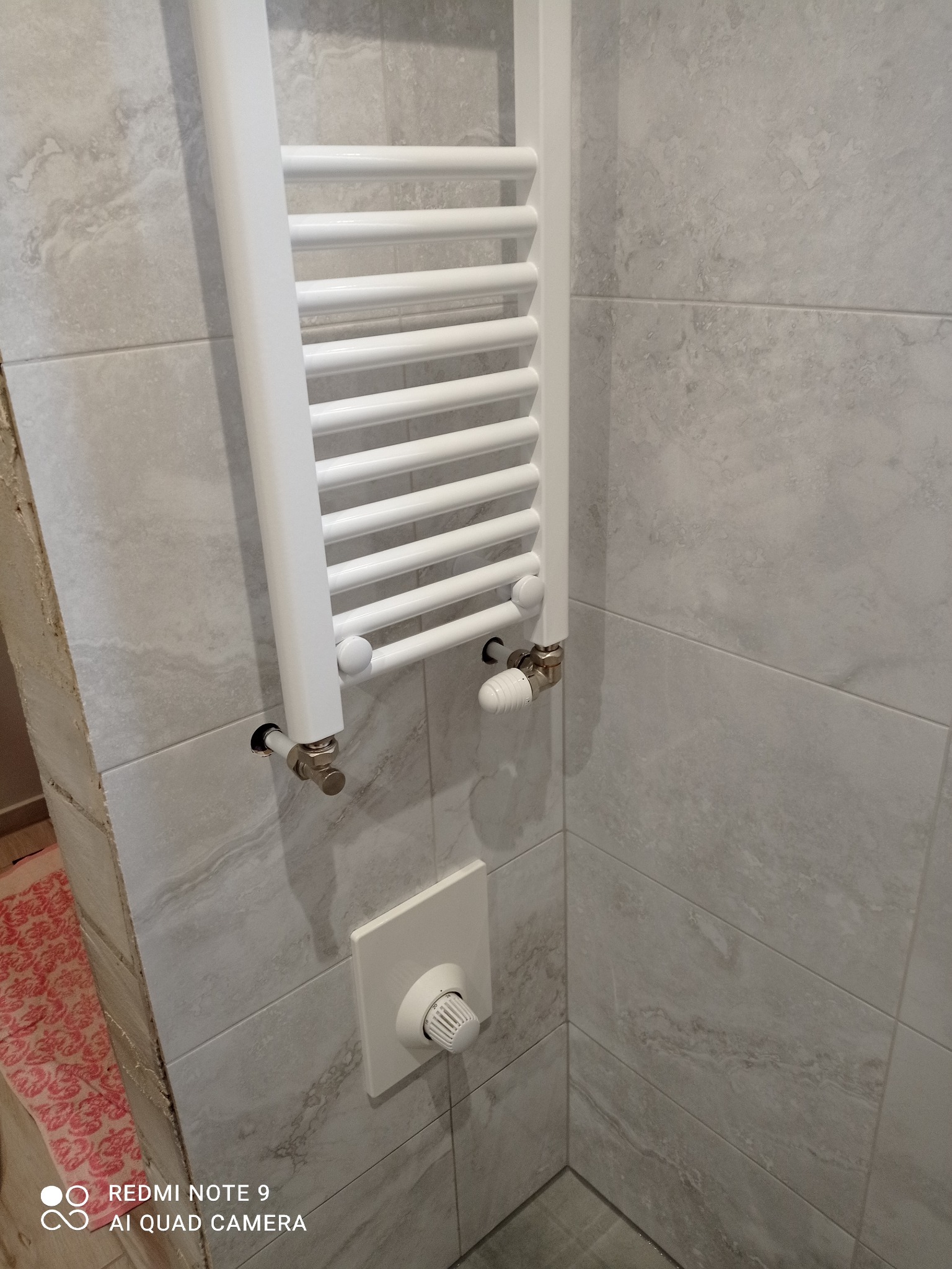 Biały grzejnik drabinkowy zamontowany na szarej, marmurkowej ścianie z płytek w łazience. Widoczny termostat i zawory. Minimalistyczny design.