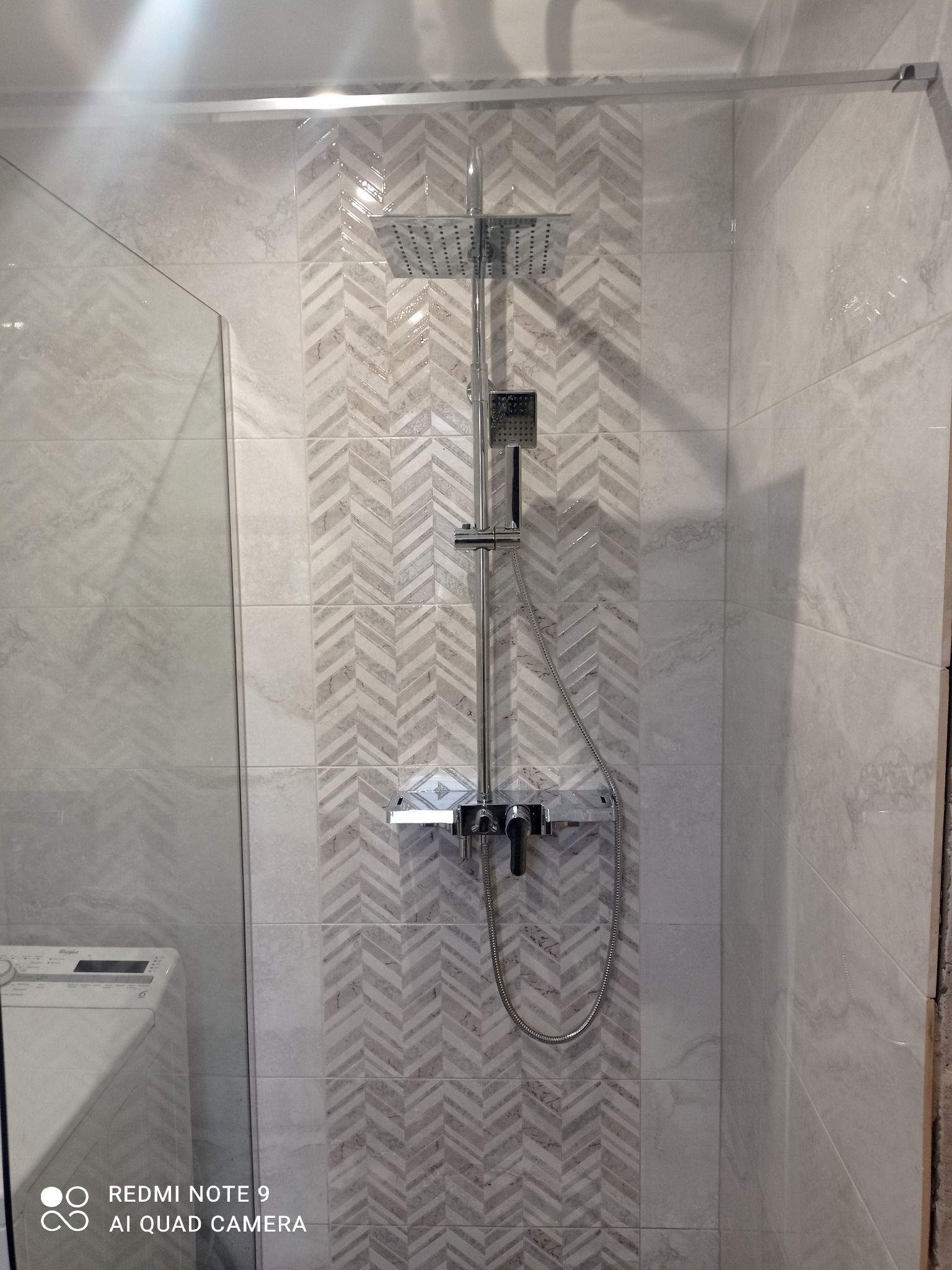 Nowoczesna kabina prysznicowa z deszczownicą i szklaną ścianką. Na ścianie płytki ułożone w jodełkę. W tle widoczna pralka. Minimalistyczny design.