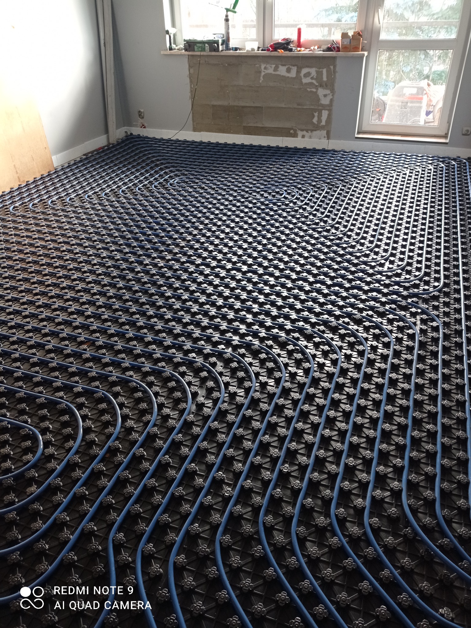 Niebieskie rurki ogrzewania podłogowego ułożone na czarnej macie w jasnym wnętrzu. Widoczne okno i fragment ściany z niedokończonym tynkiem.