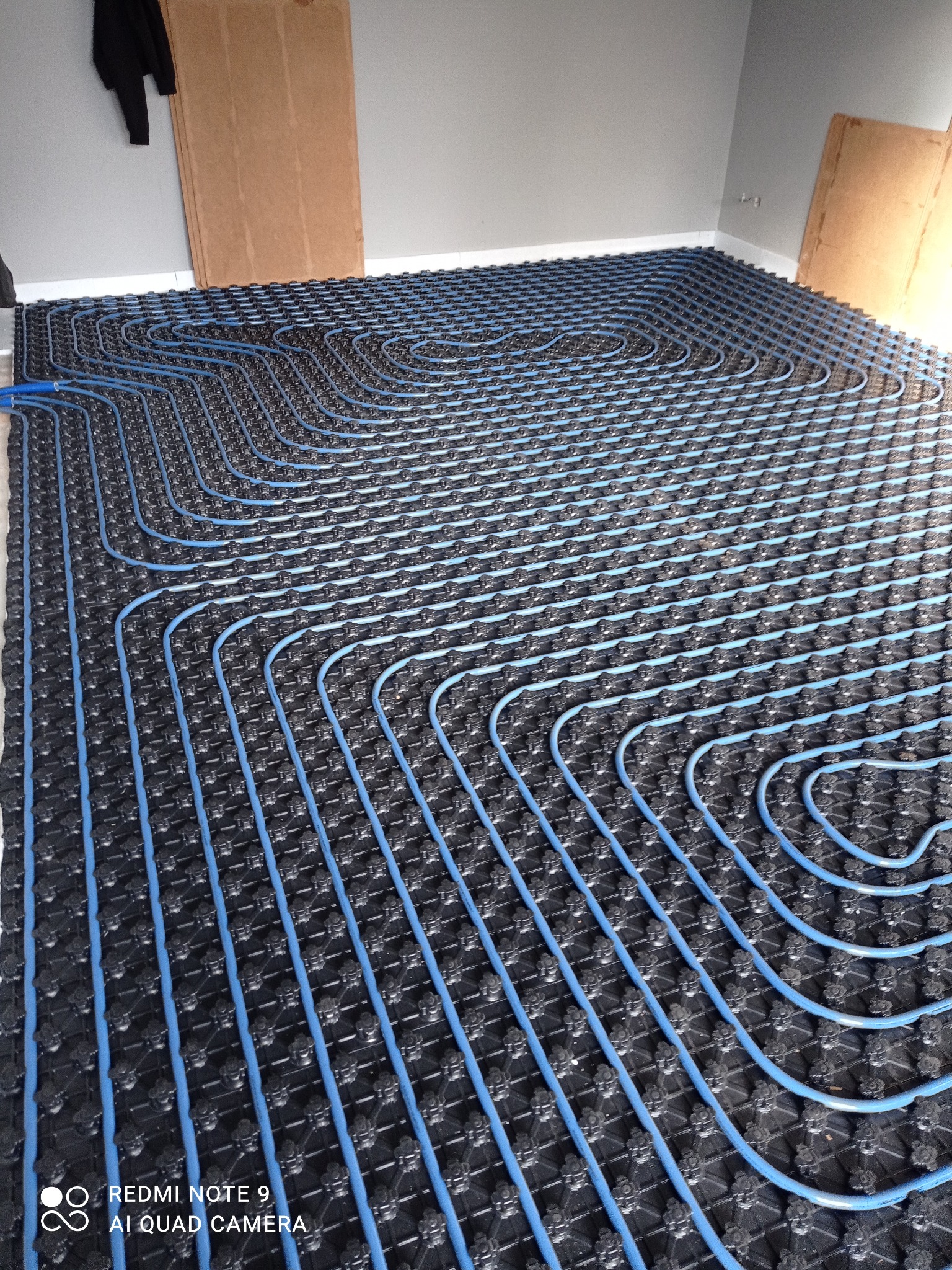 Niebieskie rurki ogrzewania podłogowego ułożone na czarnej macie izolacyjnej w szarym pomieszczeniu. Widoczne płyty kartonowo-gipsowe oparte o ścianę.
