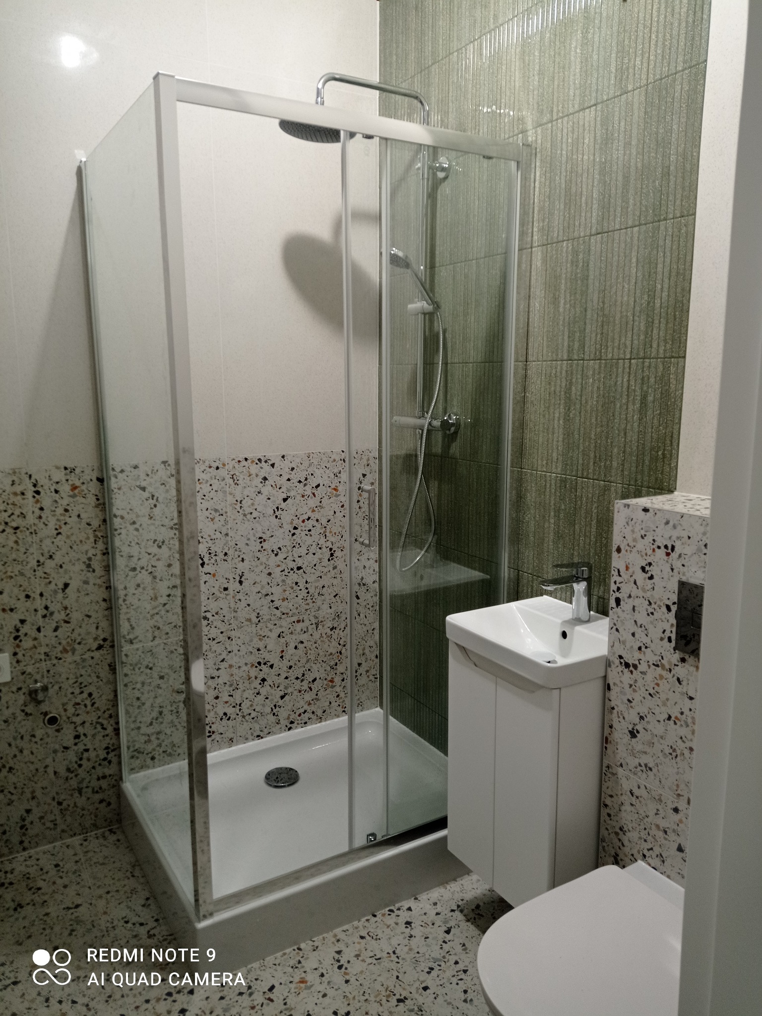 Nowoczesna łazienka po remoncie: kabina prysznicowa ze szklanymi drzwiami, umywalka z szafką i toaleta. Ściany wyłożone płytkami w dwóch odcieniach szarości.