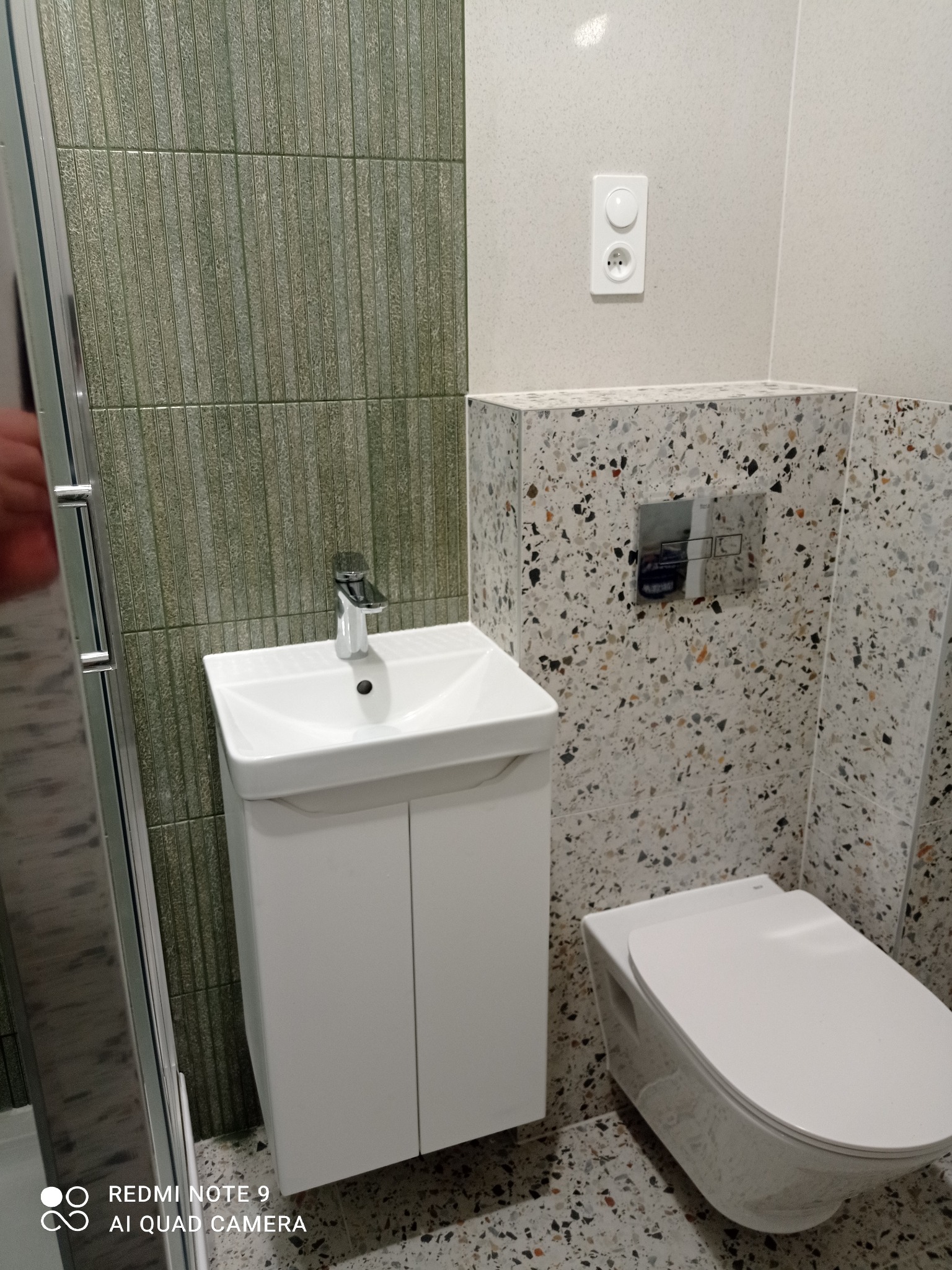 Nowoczesna łazienka po metamorfozie: biała umywalka z szafką, toaleta wisząca i mozaika na ścianach. Widoczne detale wykończenia i armatura.