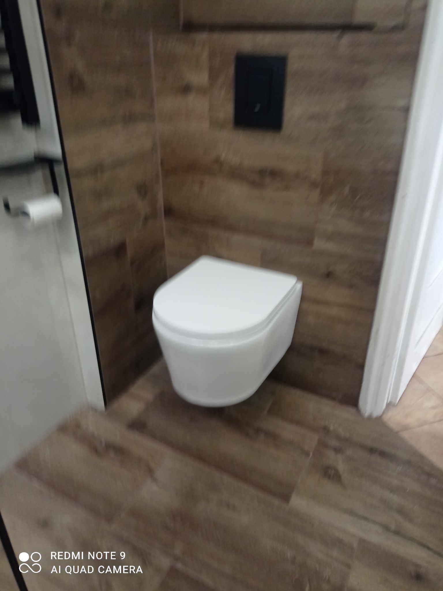 Nowoczesna toaleta wisząca w łazience wykończonej płytkami imitującymi drewno. Czarny przycisk spłuczki na ścianie. Minimalistyczny design.