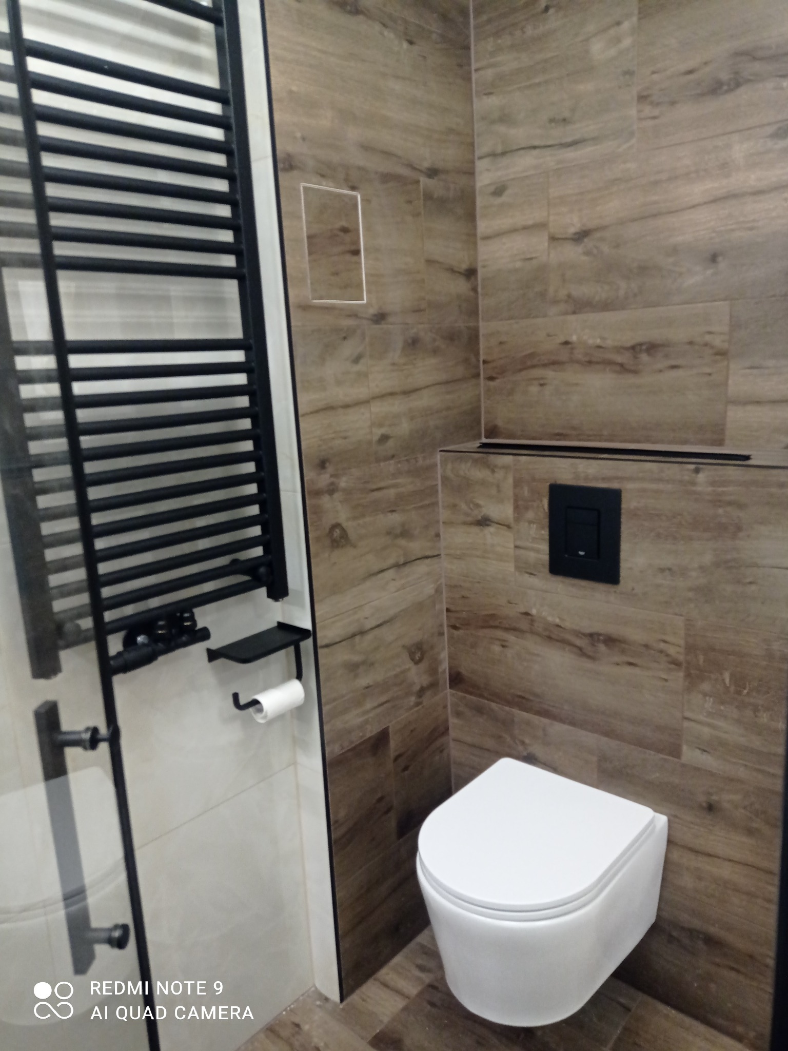 Nowoczesna łazienka z wiszącą toaletą i grzejnikiem drabinkowym na tle płytek imitujących drewno. Minimalistyczny design z czarnymi akcentami.