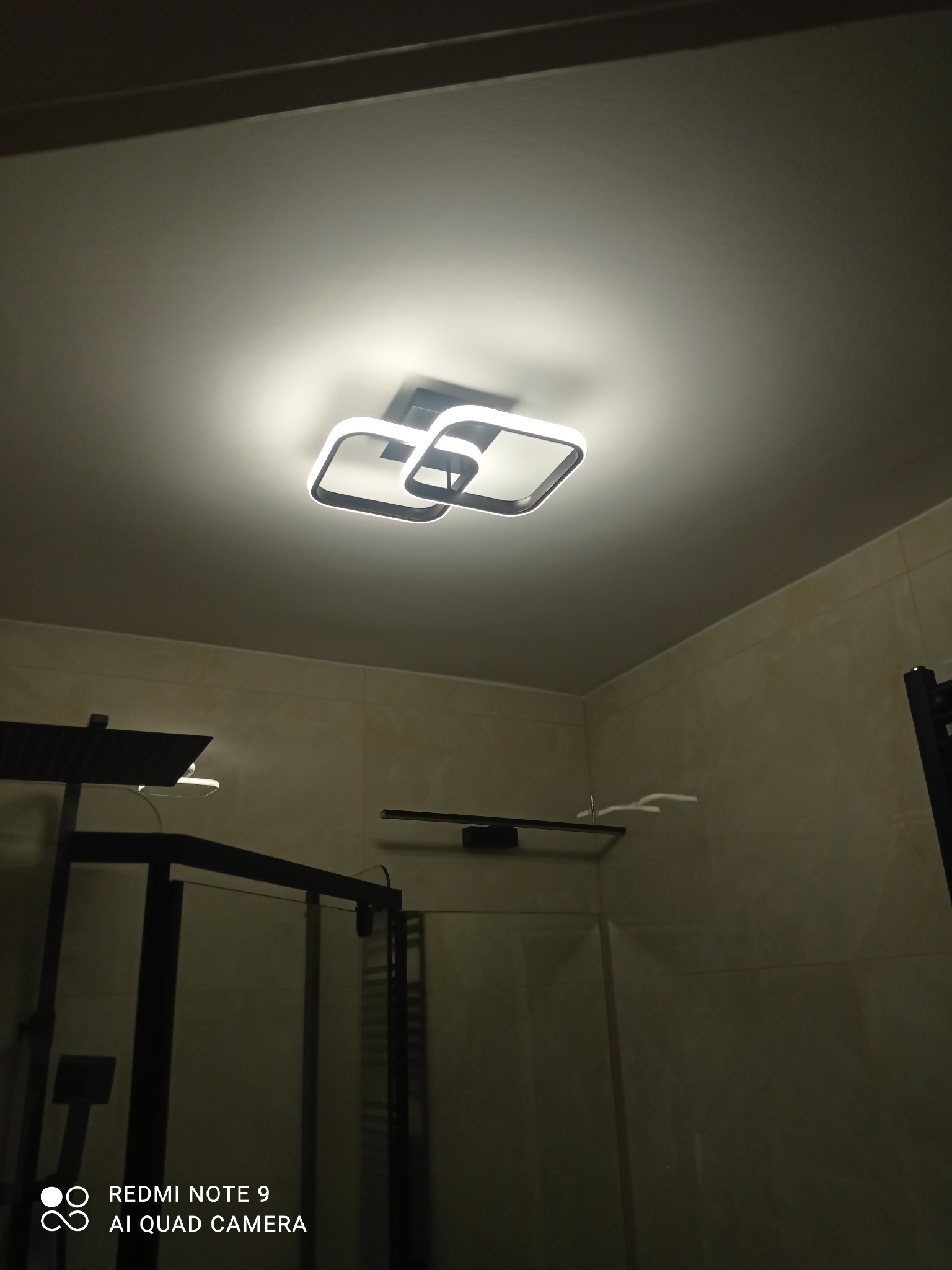 Nowoczesna lampa sufitowa LED z czarnymi obramowaniami w wykończonej łazience z beżowymi płytkami i czarną kabiną prysznicową. Minimalistyczny design.