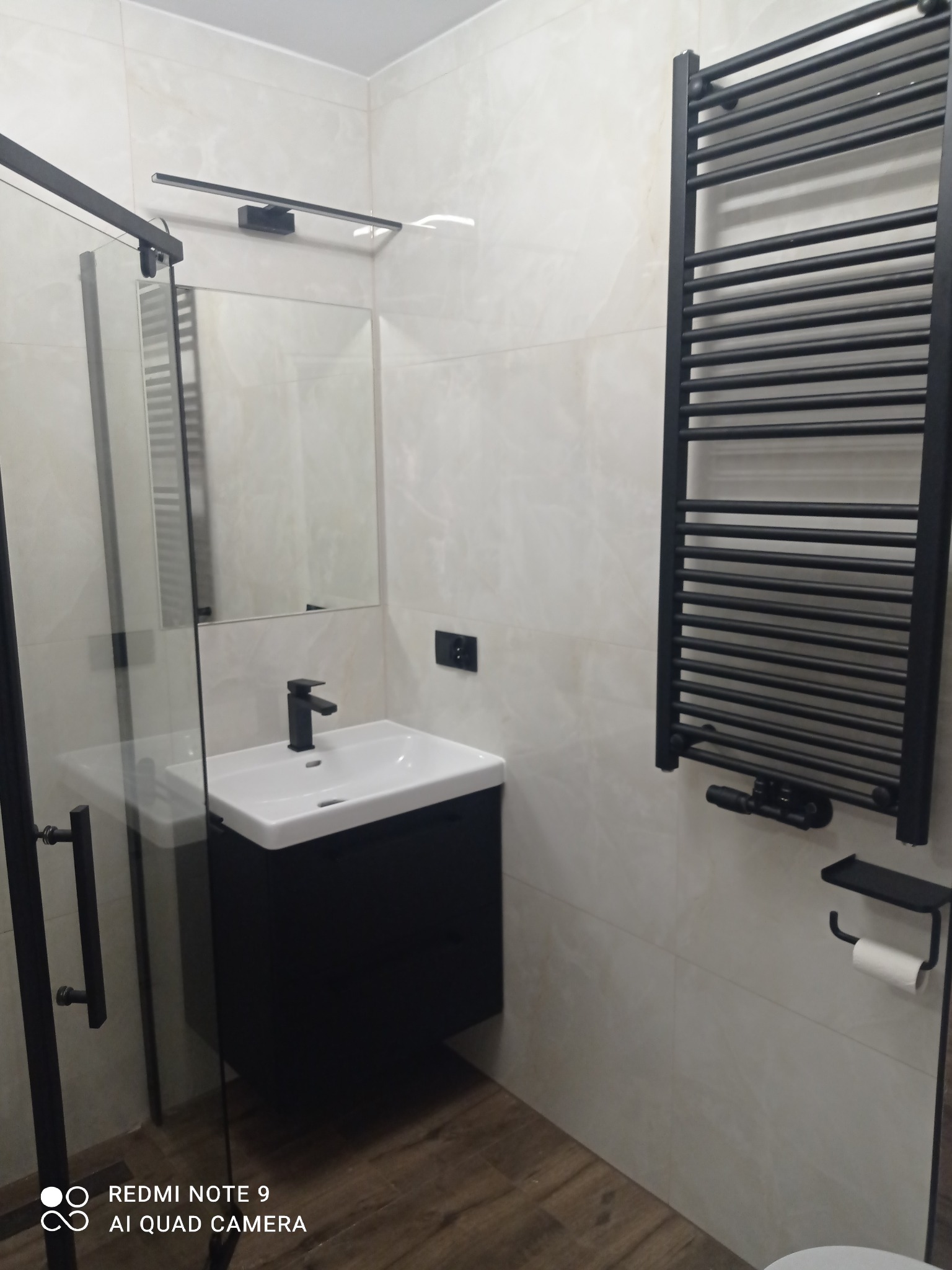 Nowoczesna łazienka z czarnymi akcentami: kabina prysznicowa, umywalka z szafką, grzejnik drabinkowy i toaletowy uchwyt na papier. Jasne płytki i drewnopodobna podłoga.