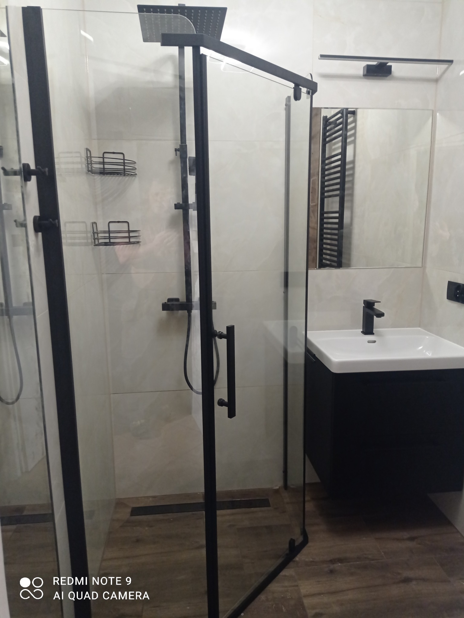 Nowoczesna kabina prysznicowa z czarnymi profilami i deszczownicą, umywalka z szafką w kolorze czarnym, jasne płytki imitujące marmur i drewnopodobna podłoga.