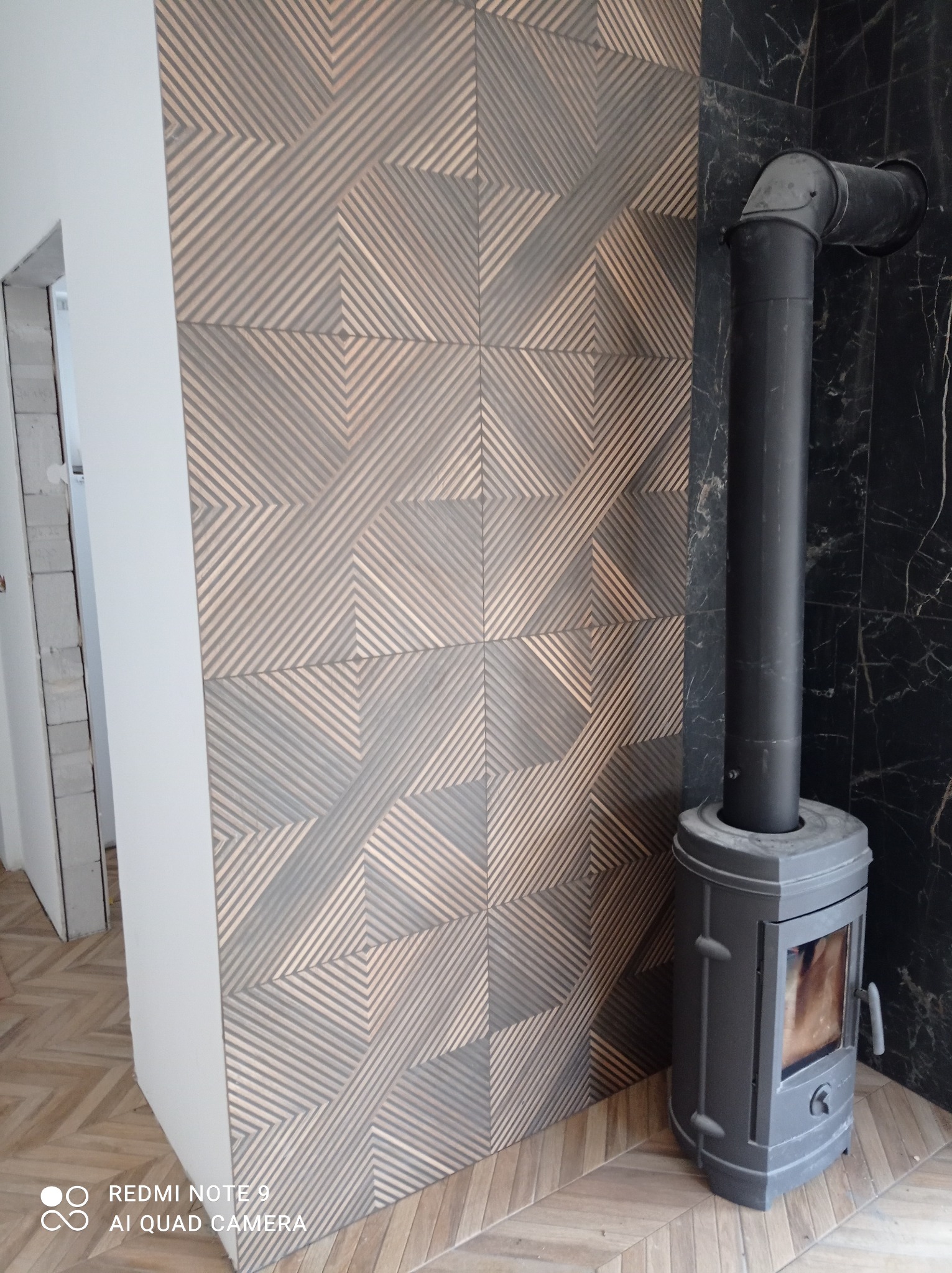 Nowoczesna aranżacja ściany z geometryczną mozaiką i czarnym marmurem, obok stoi piec kominkowy z rurą. Parkiet ułożony w jodełkę na podłodze.