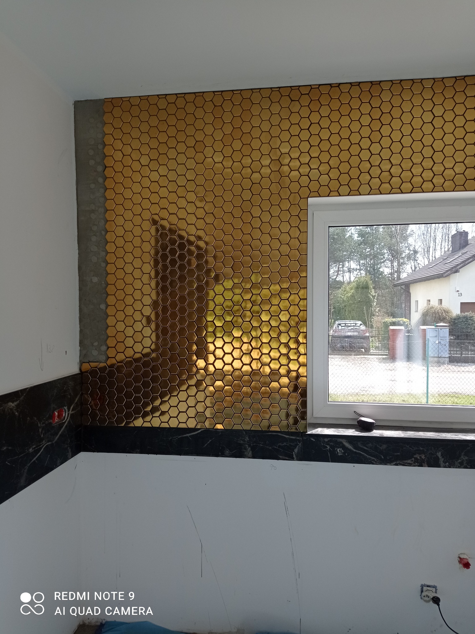 Ściana z mozaiki w kształcie złotych heksagonów, częściowo wykończona, z widocznym oknem i fragmentem wnętrza w trakcie prac wykończeniowych.