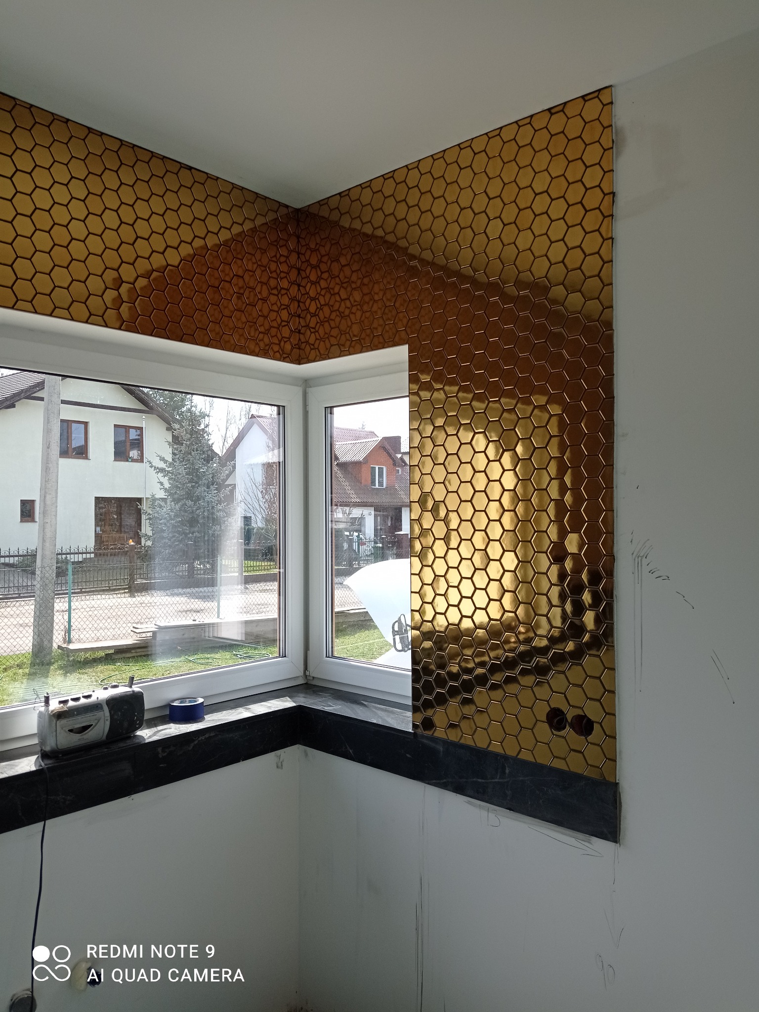 Złote, heksagonalne płytki ułożone na ścianie w kuchni, widoczne przy oknie. Częściowo wykończona ściana, czarny blat, radio na parapecie. Widok na domy za oknem.