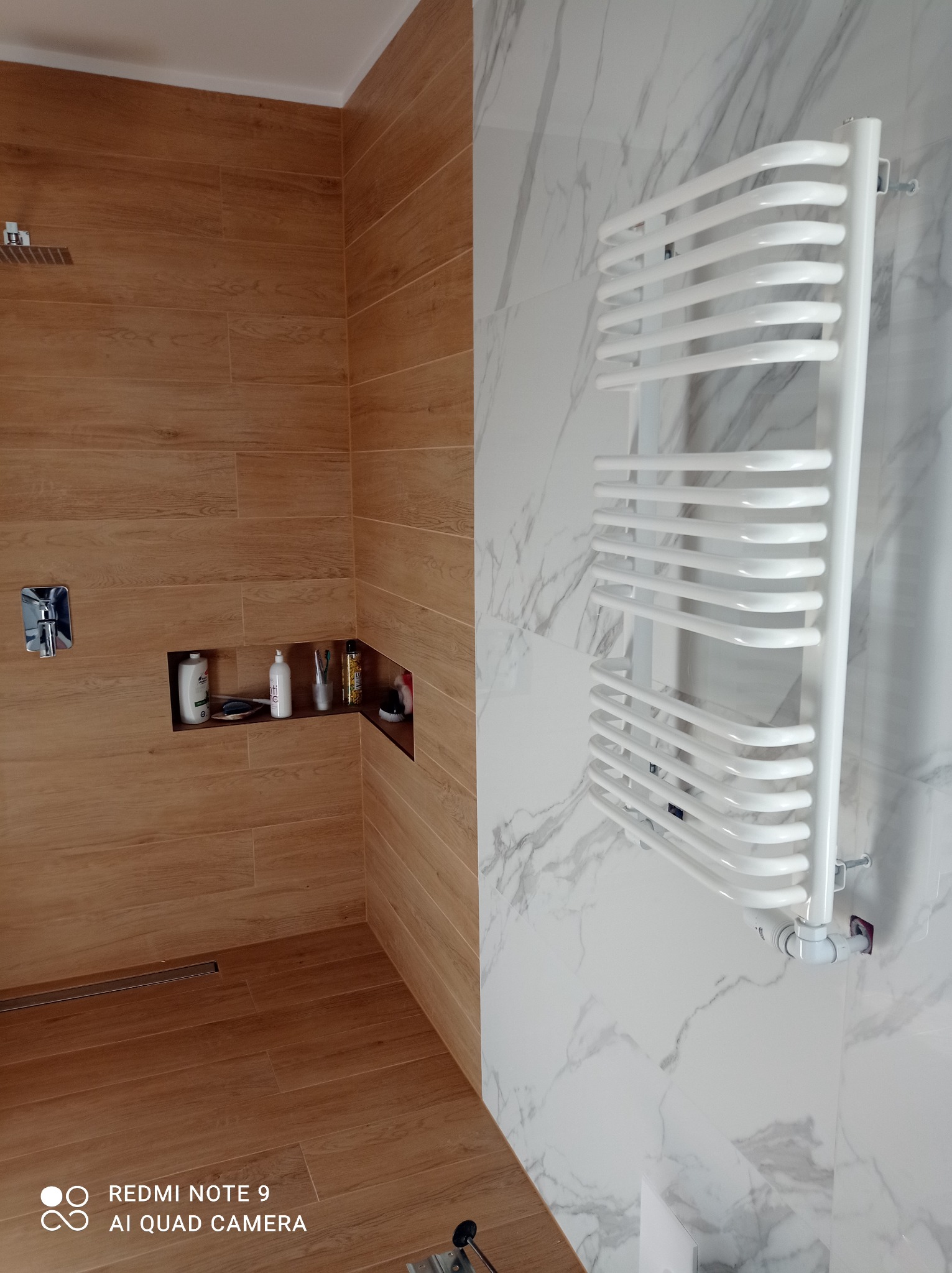 Nowoczesna łazienka z drewnopodobnymi płytkami, kabiną prysznicową typu walk-in i białym grzejnikiem drabinkowym na marmurowej ścianie.