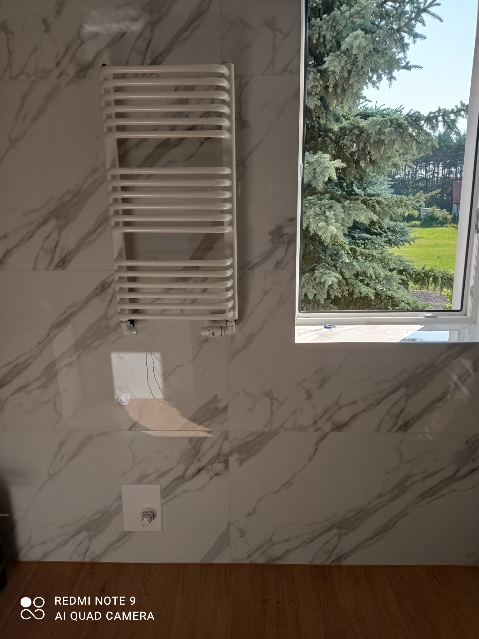 Biały grzejnik drabinkowy na ścianie z płytek imitujących marmur, obok okno z widokiem na zieleń. Minimalistyczny design łazienki z akcentem natury.