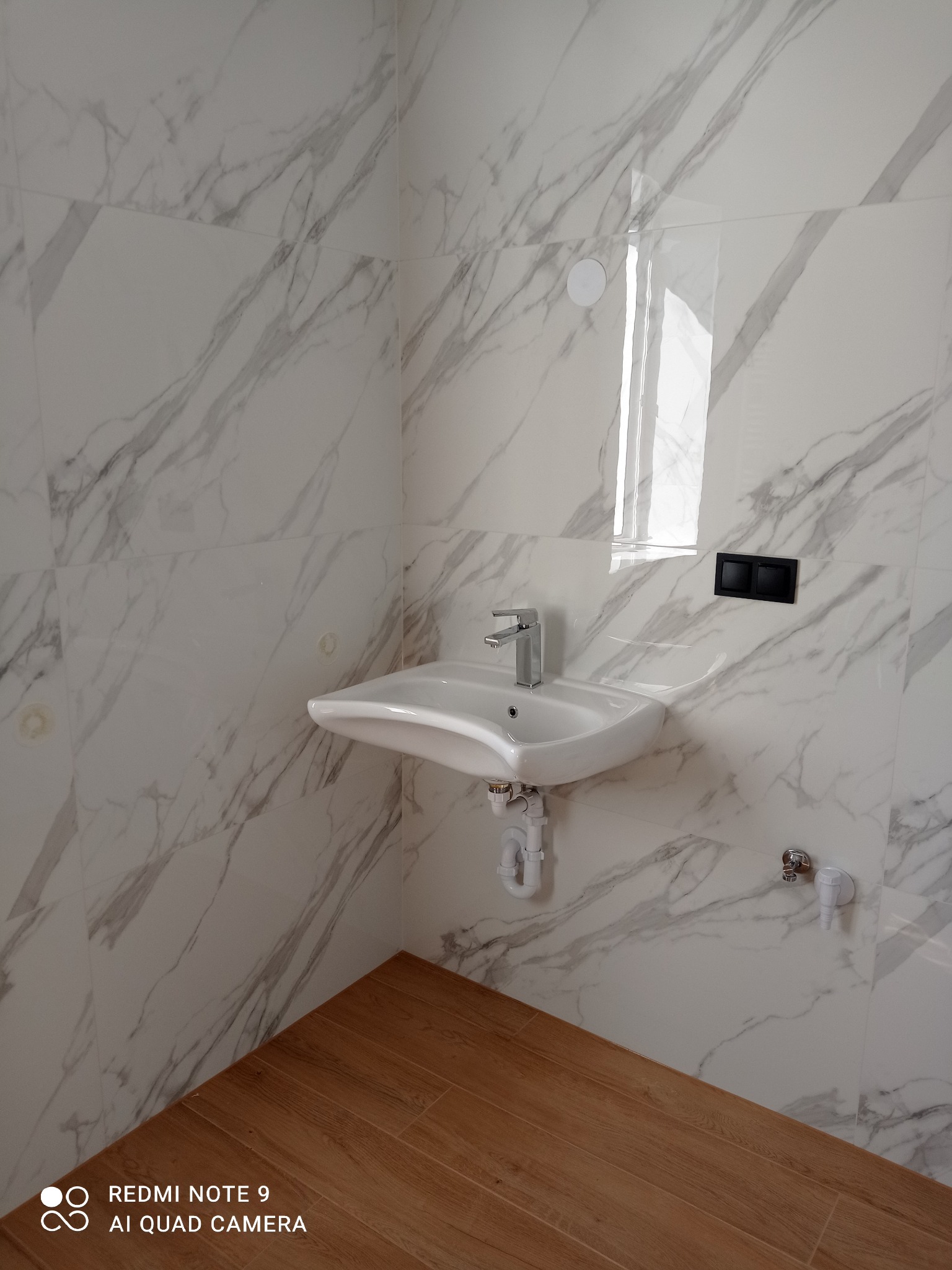 Biała umywalka z chromowaną baterią na tle marmurowych płytek i drewnopodobnej podłogi. Nowoczesna armatura łazienkowa po montażu.