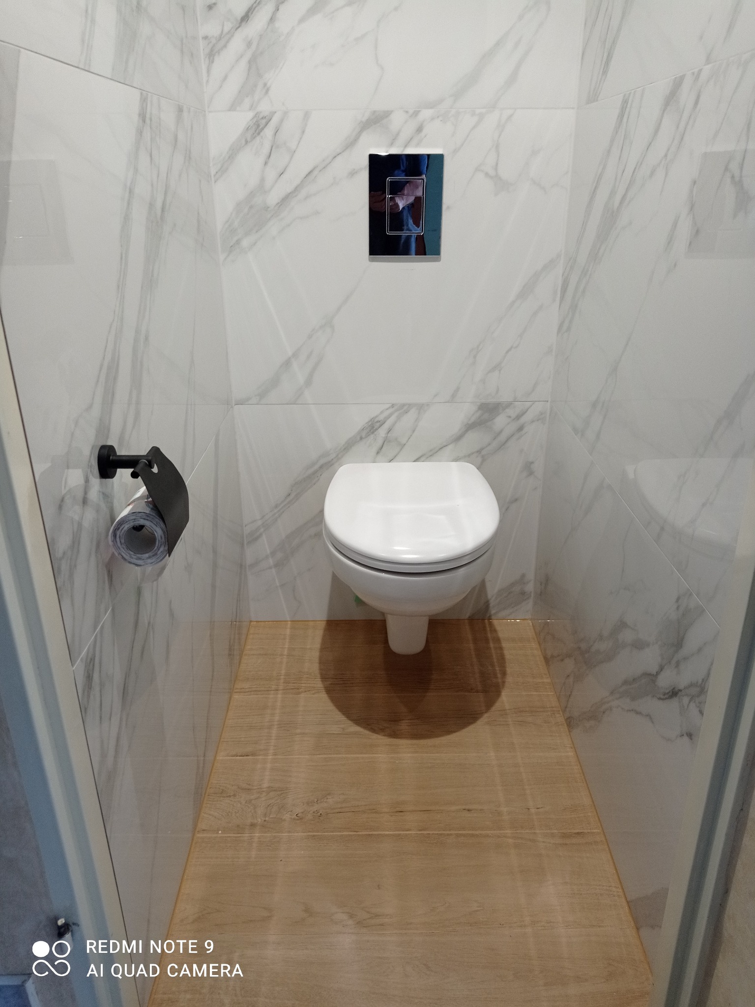 Nowoczesna toaleta podwieszana w wykończonej łazience. Białe płytki imitujące marmur na ścianach, drewnopodobna podłoga, czarny uchwyt na papier toaletowy.