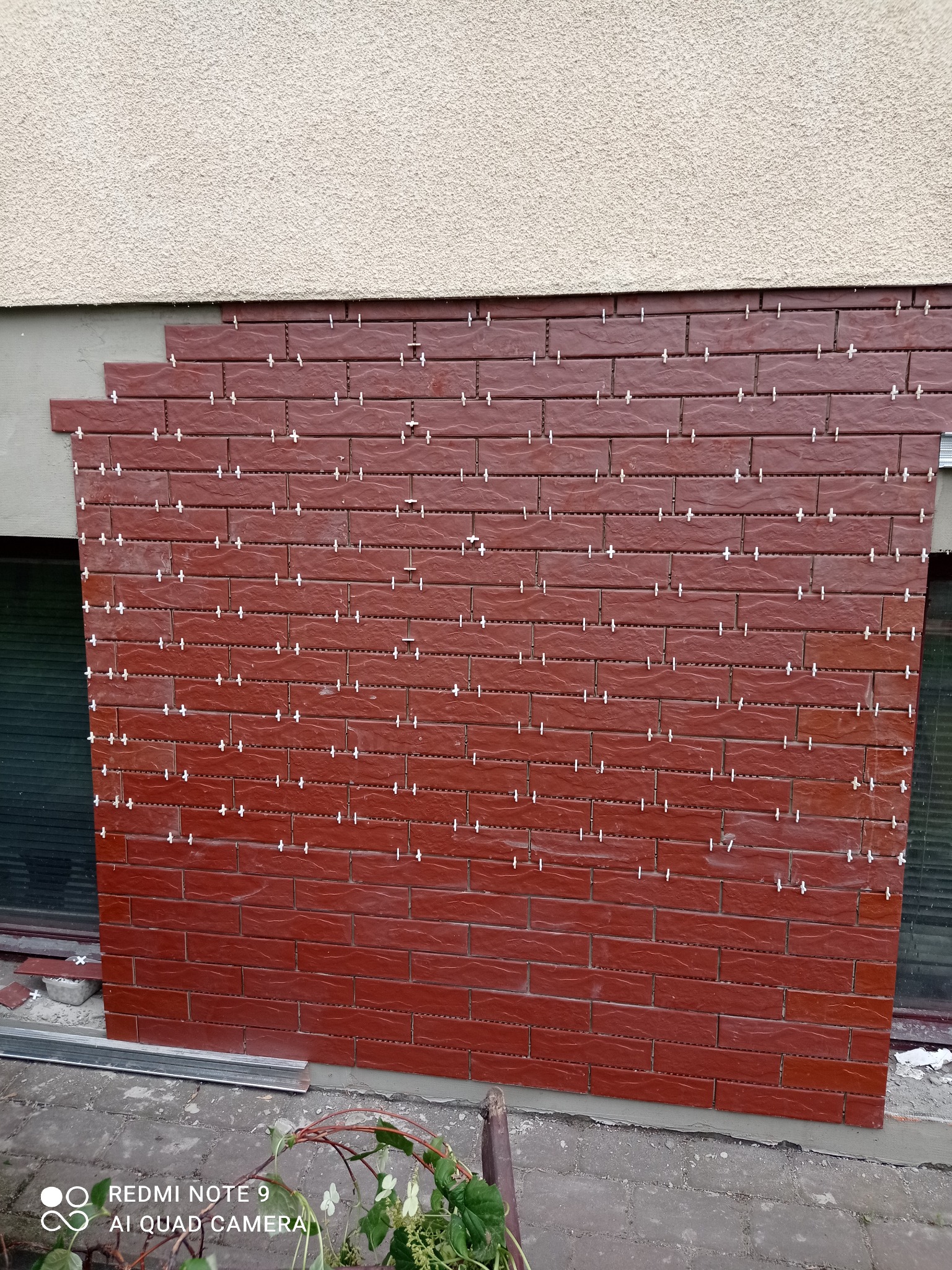 Ceglana okładzina elewacyjna w trakcie montażu, z widocznymi krzyżykami dystansowymi. Częściowo wykończona ściana zewnętrzna budynku.