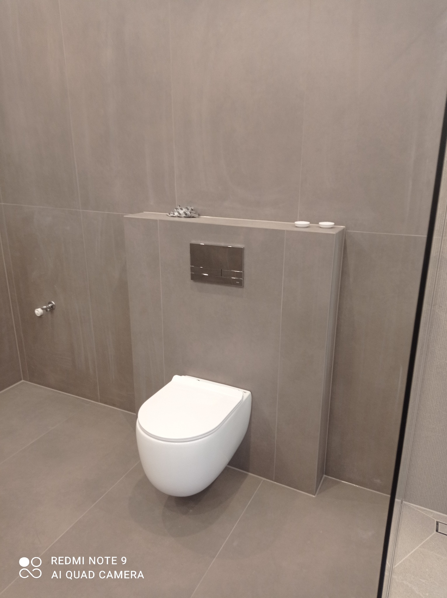 Podwieszana toaleta w minimalistycznej łazience, wykończonej szarymi płytkami. Widoczny metalowy panel spłukujący i akcesoria łazienkowe na półce.