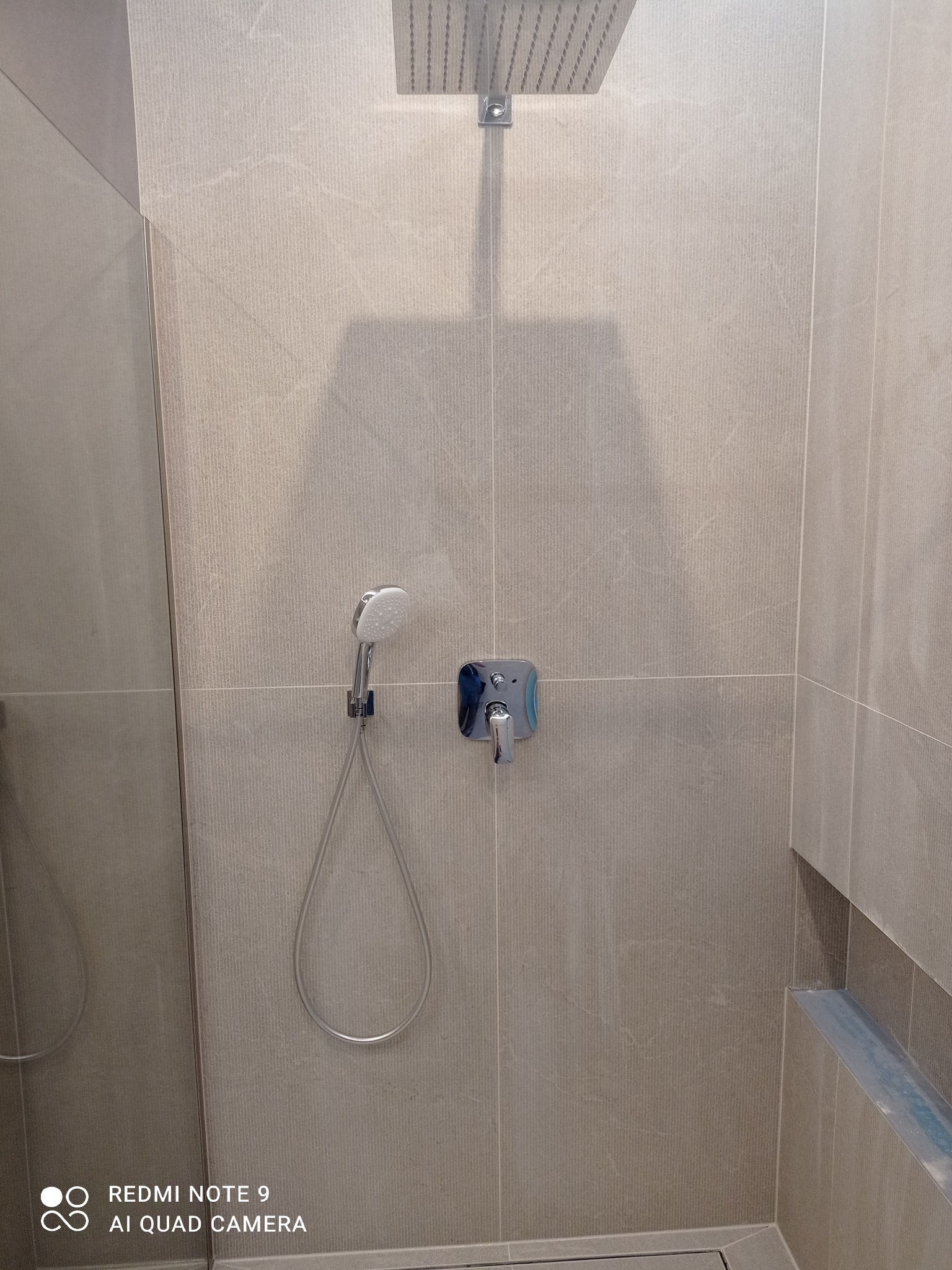 Wykończona kabina prysznicowa z deszczownicą i baterią, wyłożona beżowymi płytkami imitującymi kamień. Widoczny odpływ liniowy i półka wnękowa.