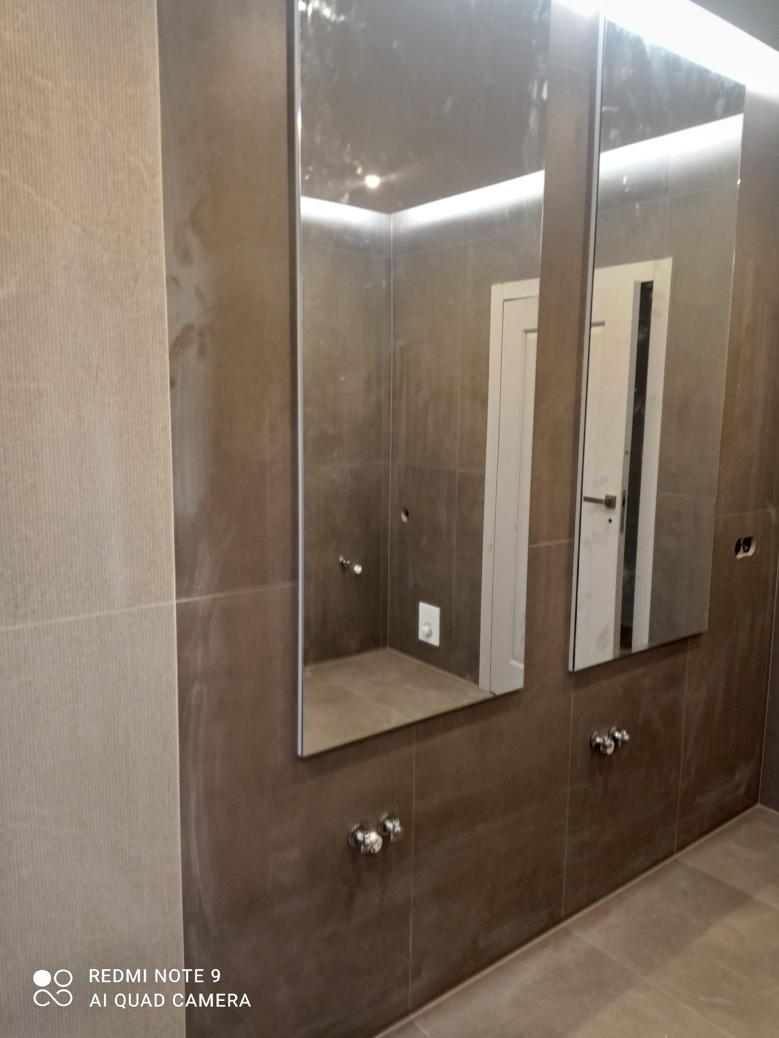 Wykończona łazienka z dwoma lustrami z podświetleniem LED, brązowe płytki na ścianach i podłodze, widoczne baterie umywalkowe. Minimalistyczny design.