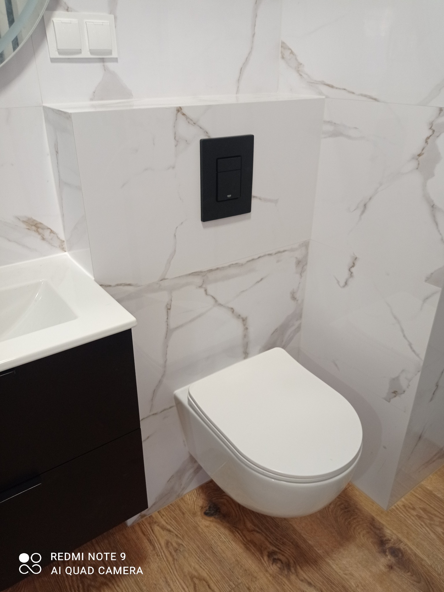 Nowoczesna łazienka z podwieszaną toaletą, czarnym przyciskiem spłukującym i umywalką z szafką. Białe płytki imitujące marmur na ścianach i drewnopodobna podłoga.