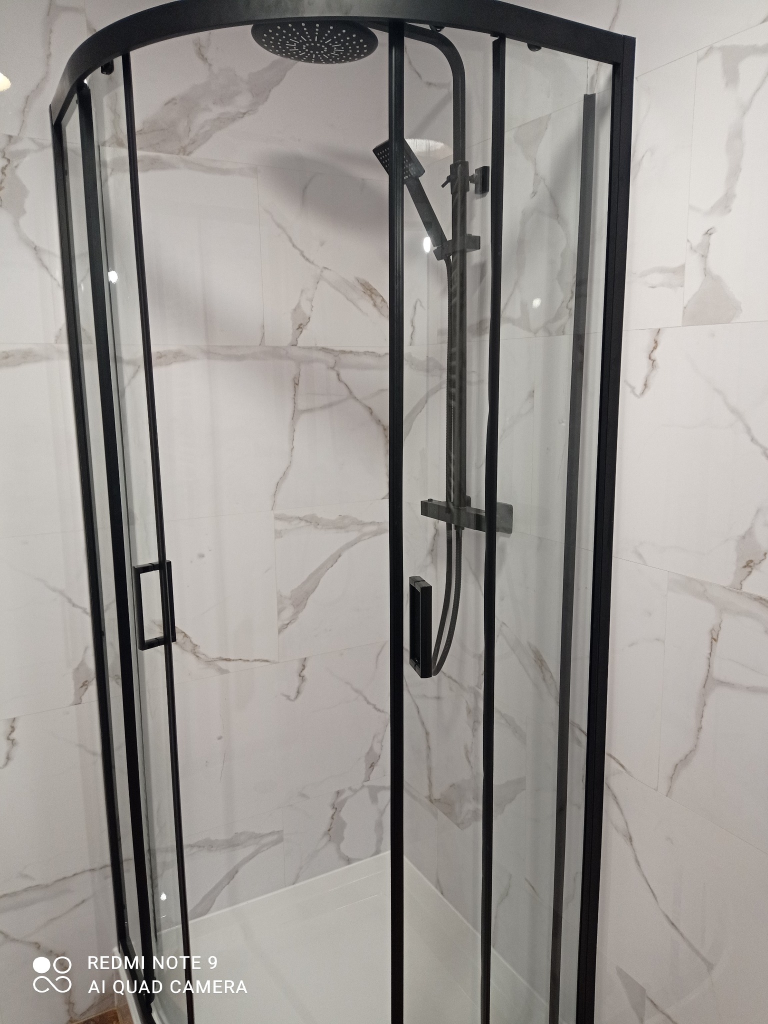 Kabina prysznicowa z czarnymi profilami na tle marmurowych płytek. Nowoczesny design, widoczny zestaw natryskowy. Wykończona łazienka w jasnych barwach.