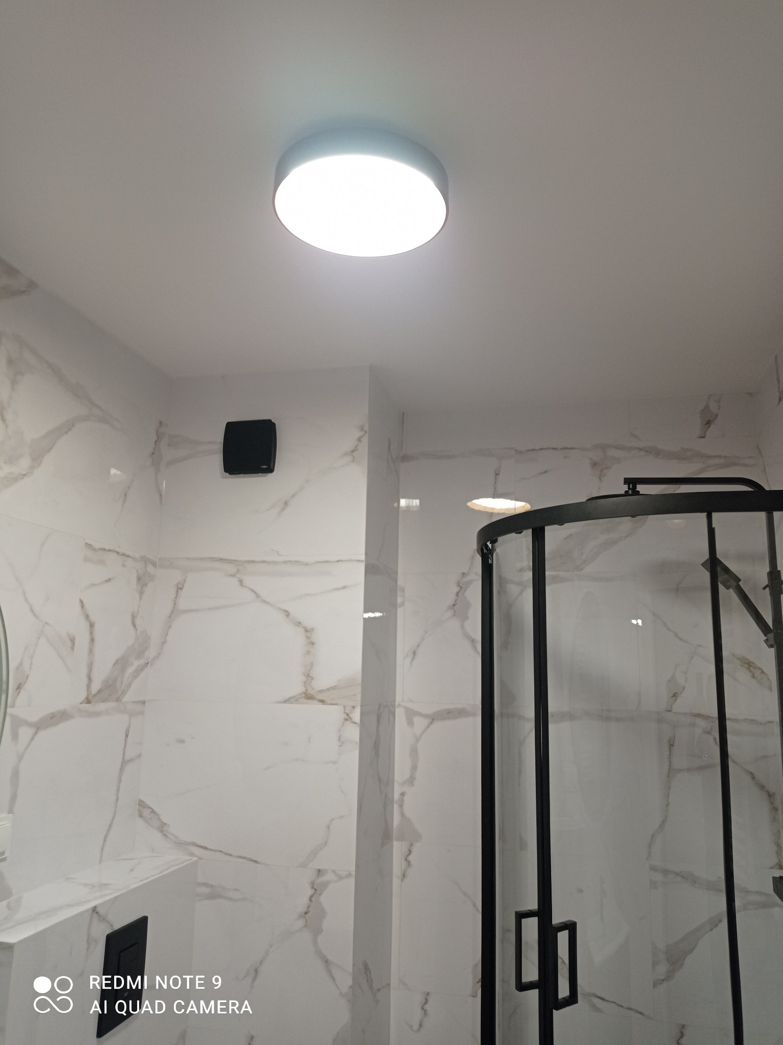 Biała łazienka z marmurowymi płytkami, czarną kabiną prysznicową i okrągłą lampą sufitową. Minimalistyczny design, nowoczesne wykończenie.