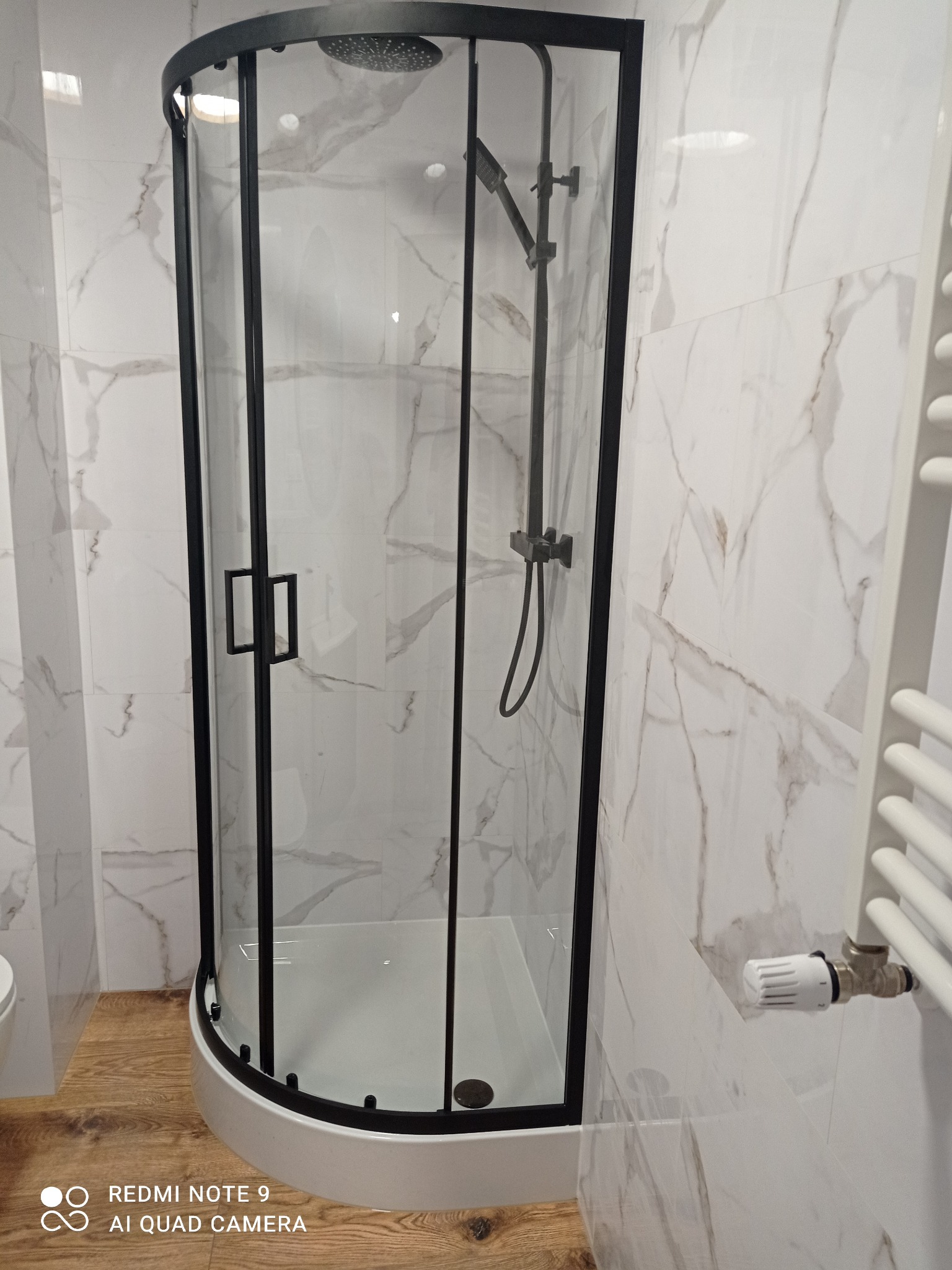 Nowoczesna kabina prysznicowa z czarnymi profilami na tle marmurowych płytek i drewnopodobnej podłogi. Widoczny grzejnik drabinkowy i termostat.