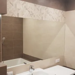 Ukończona łazienka w beżowo-brązowej kolorystyce z dużym lustrem, widoczna bateria umywalkowa, fragment wanny i pralki, nad lustrem dekoracyjny pas z wzorzystych płytek.