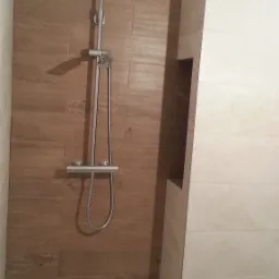 Wykończona kabina prysznicowa z brązowymi płytkami imitującymi drewno i chromowaną armaturą, widoczna wnęka w ścianie obłożonej jasnymi płytkami.