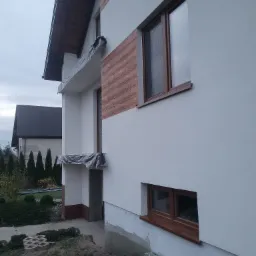 Fragment elewacji domu w Skarżysko-Kamienna z widoczną świeżo położoną białą strukturą tynku oraz imitacją drewnianej okładziny, okna z brązowymi ramami, zabezpieczone elementy przed zabrudzeniem.