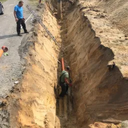 Głęboki wykop w ziemi z widoczną instalacją rur, dwóch pracowników i drabiną; teren budowy z pryzmami ziemi.