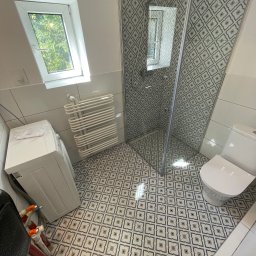 Wg home invest - Ukończona łazienka z kabiną prysznicową, toaletą, grzejnikiem drabinkowym i pralką, wykończona płytkami podłogowymi w geometryczny wzór. Widoczny fragment buta w rogu kadru sugeruje obecność osoby...