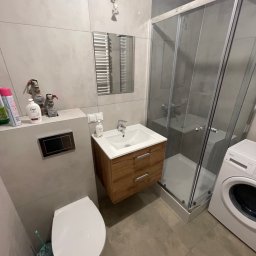 Wg home invest - Nowoczesna łazienka z kabiną prysznicową, umywalką z drewnianą szafką, toaletą i pralką. Szare płytki na ścianach i podłodze. Widoczny grzejnik drabinkowy i wentylator. Uwagę przyciąga nietypowe...