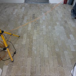 Wg home invest - Ułożone płytki podłogowe w trakcie fugowania, widoczne krzyżyki dystansowe, oświetlenie budowlane na trójnogu i przewody elektryczne.