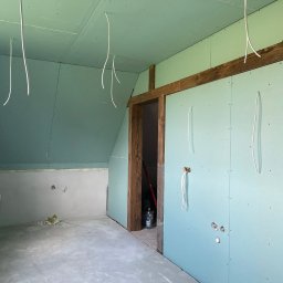 Wg home invest - Poddasze w trakcie prac wykończeniowych: zamontowane płyty karton-gips, widoczne kable zwisające z sufitu, surowe drewniane ramy drzwiowe i fragment ściany przygotowanej do dalszych prac.
