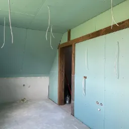 Poddasze w trakcie prac wykończeniowych: zamontowane płyty karton-gips, widoczne kable zwisające z sufitu, surowe drewniane ramy drzwiowe i fragment ściany przygotowanej do dalszych prac.