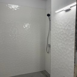 Wg home invest - Nowoczesna kabina prysznicowa z białymi, trójwymiarowymi płytkami, szarymi płytkami podłogowymi z odpływem liniowym i chromowaną armaturą, oświetlona listwą LED, w trakcie remontu.
