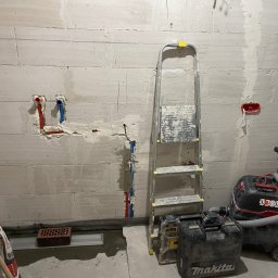 Wg home invest - Ściana z betonu komórkowego w trakcie przygotowywania do wykończenia, widoczne instalacje wodne z rurami w kolorze czerwonym i niebieskim, drabina aluminiowa, odkurzacz przemysłowy i skrzynki...
