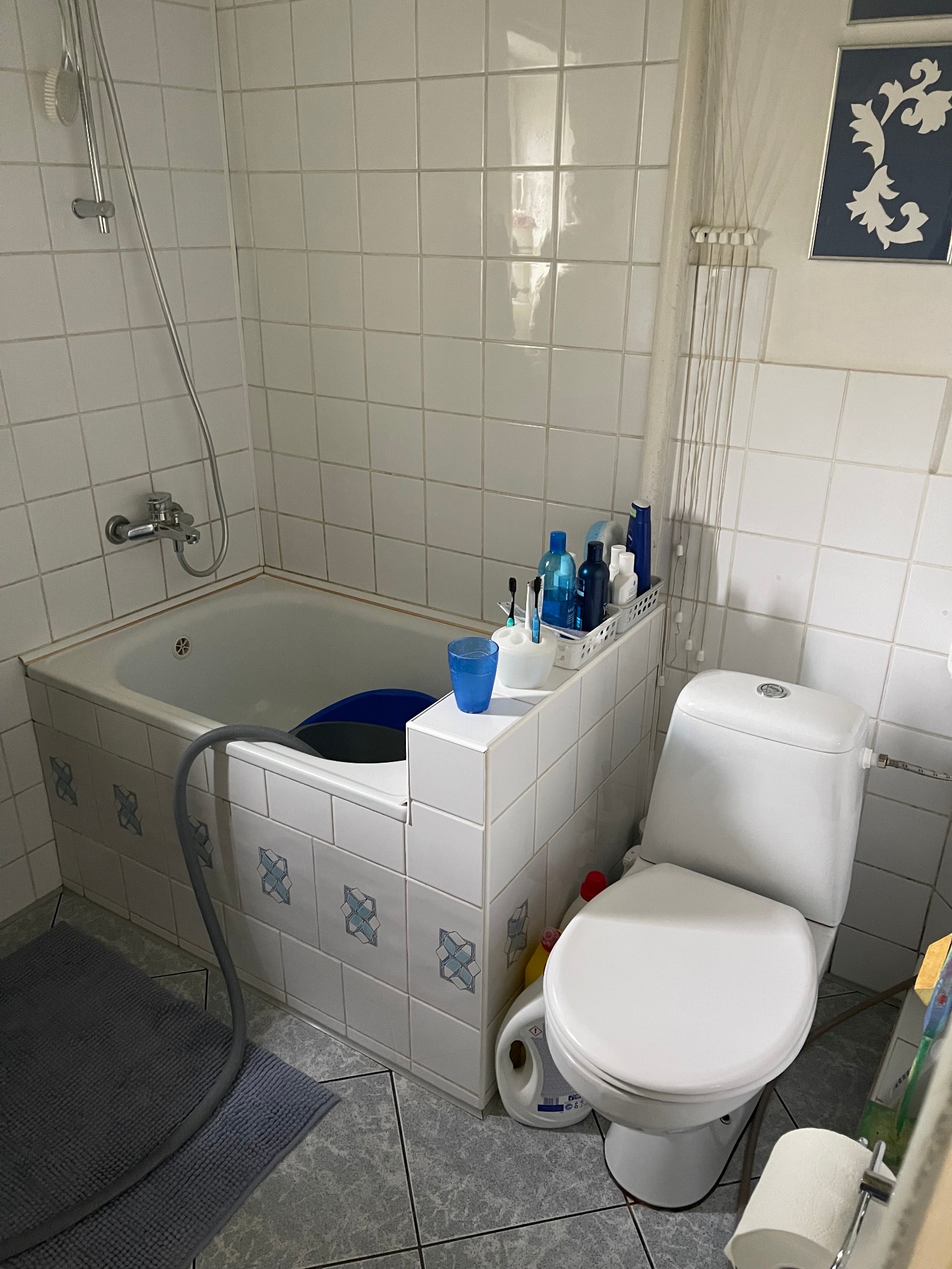 Standardowa łazienka w bloku z wanną obudowaną kafelkami z niebieskim wzorem, toaleta z pojemnikiem na środki czystości obok, szary dywanik i białe kafelki na ścianach.