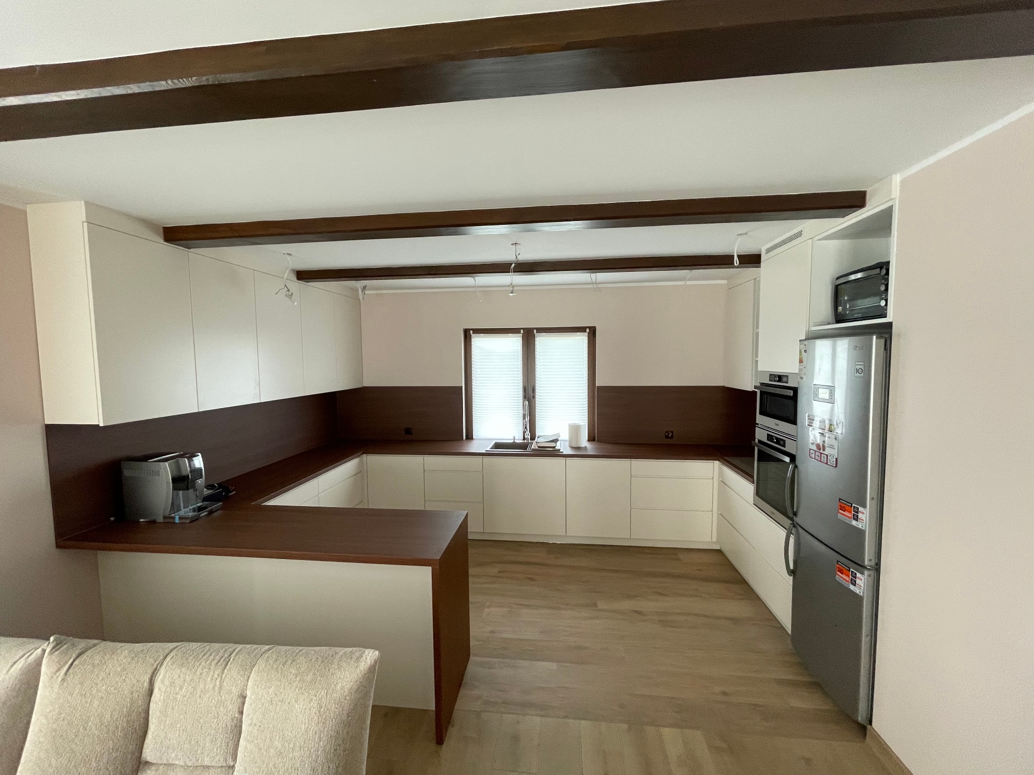 Nowoczesna kuchnia w bieli i brązie z drewnianymi belkami sufitowymi, widok na zabudowę kuchenną, sprzęt AGD i część salonu z beżową sofą.