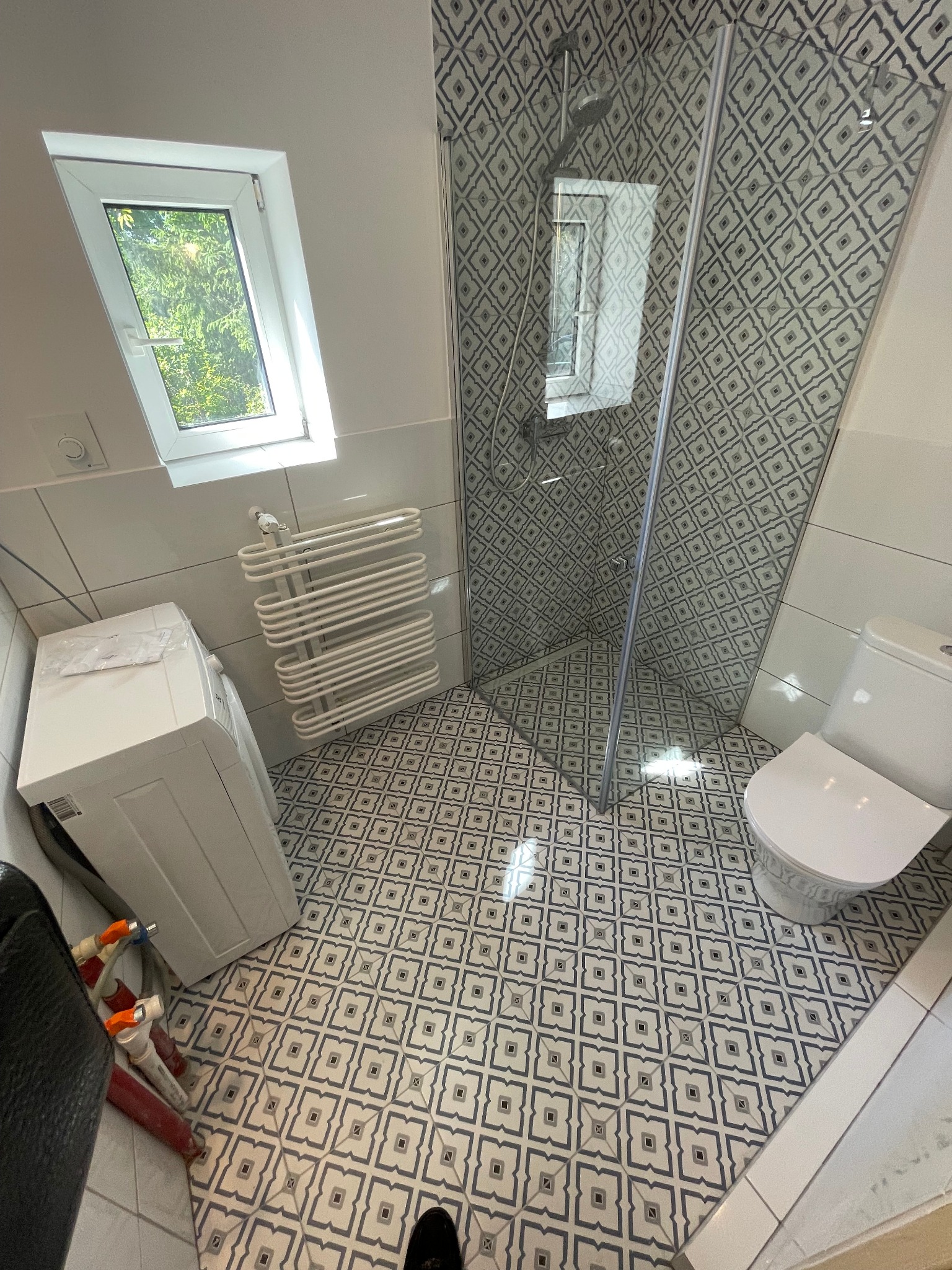 Ukończona łazienka z kabiną prysznicową, toaletą, grzejnikiem drabinkowym i pralką, wykończona płytkami podłogowymi w geometryczny wzór. Widoczny fragment buta w rogu kadru sugeruje obecność osoby...