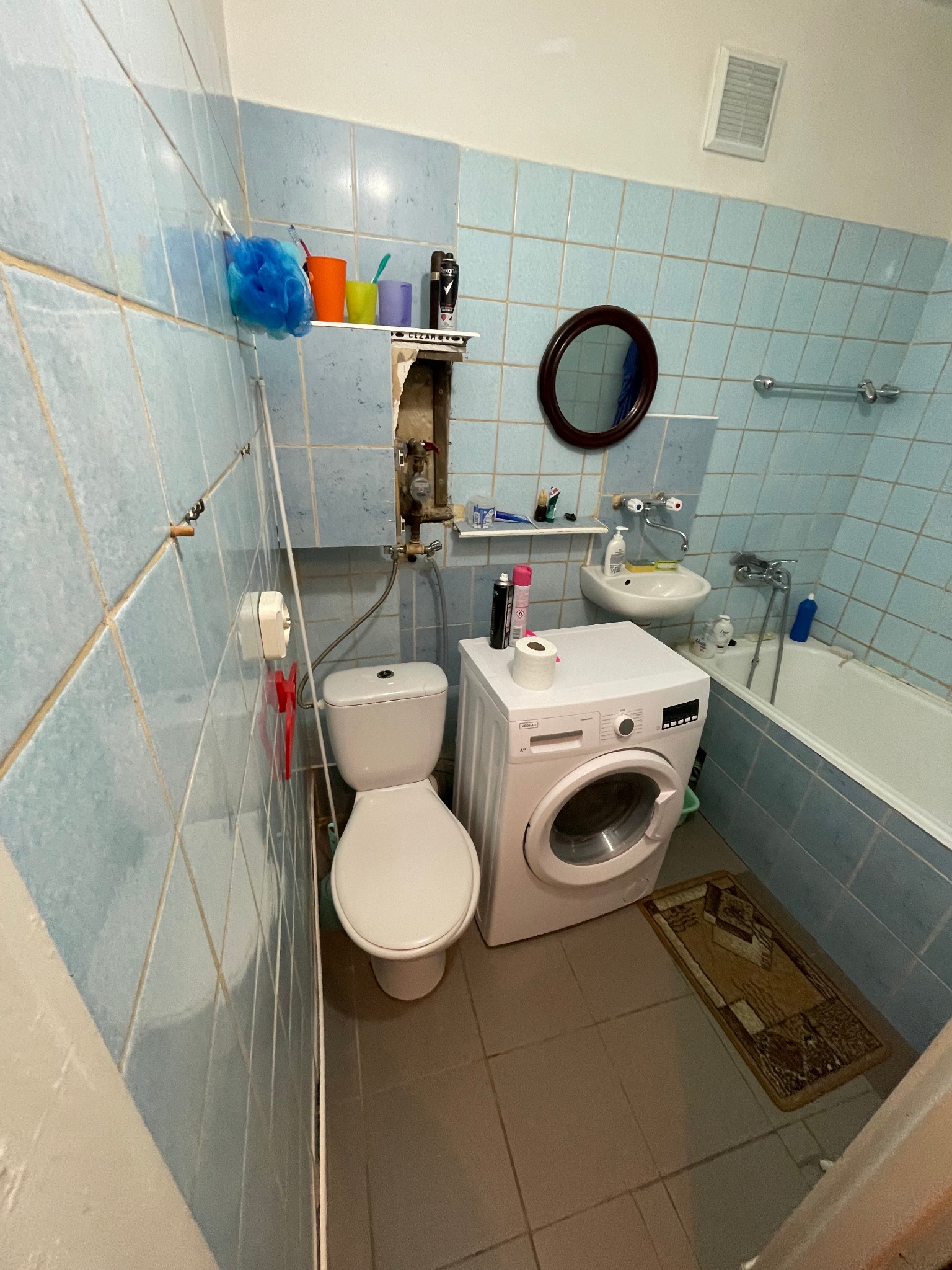 Niebieska łazienka w Sokółce z pralką, toaletą i wanną, gdzie widać odkryte rury za toaletą.