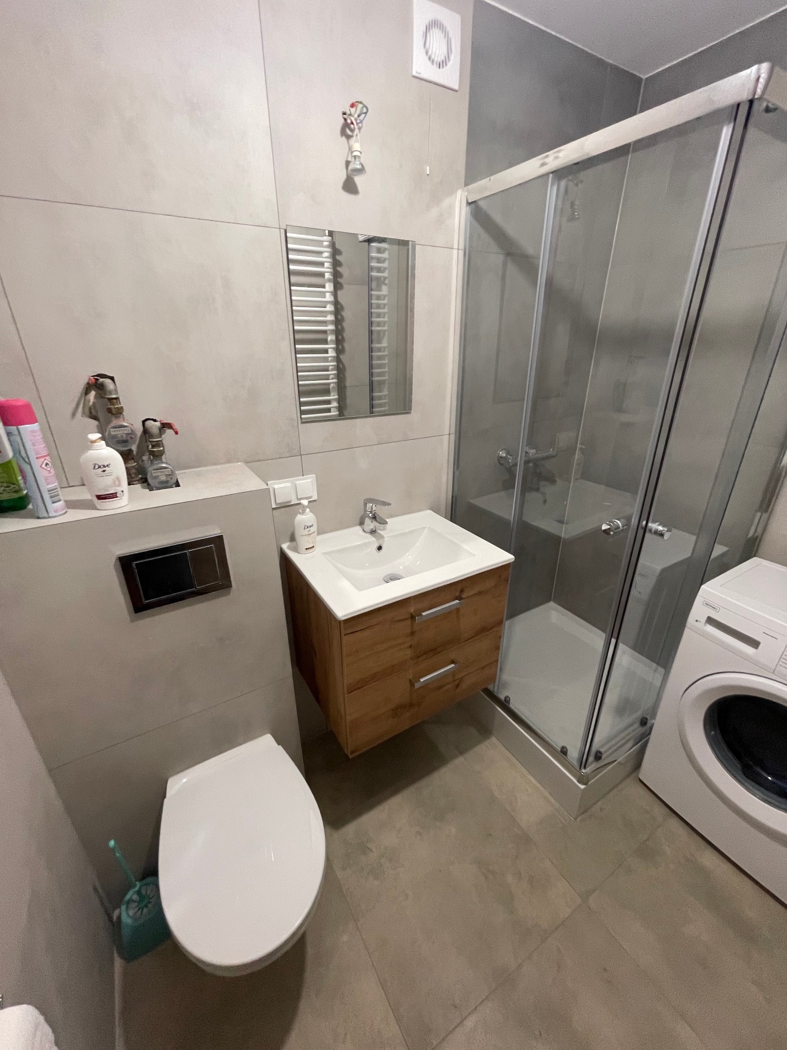 Nowoczesna łazienka z kabiną prysznicową, umywalką z drewnianą szafką, toaletą i pralką. Szare płytki na ścianach i podłodze. Widoczny grzejnik drabinkowy i wentylator. Uwagę przyciąga nietypowe...