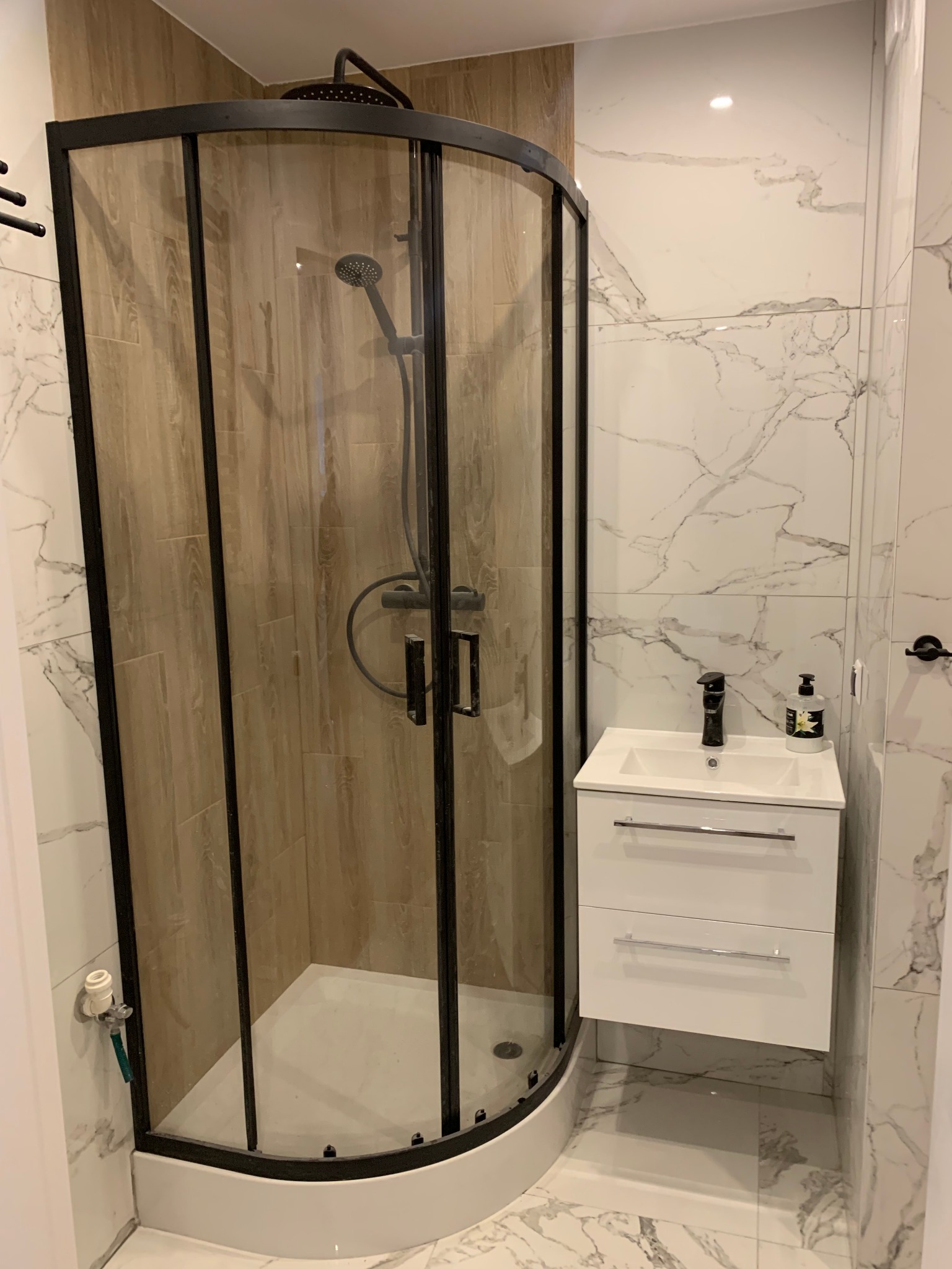 Nowoczesna łazienka z kabiną prysznicową typu walk-in z czarnymi profilami, białą umywalką z szafką i marmurowymi płytkami na ścianach i podłodze.