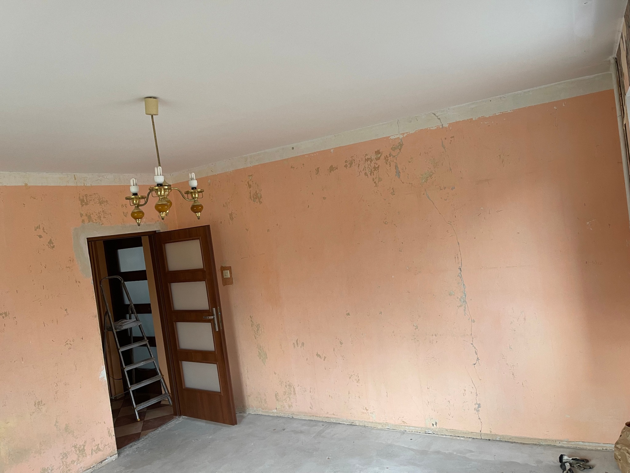 Pokój w trakcie przygotowania do malowania: ściany w kolorze brzoskwini z widocznymi ubytkami i resztkami starej farby, drabina oparta o futrynę drzwi, widoczny fragment sufitu z lampą. Podłoga...