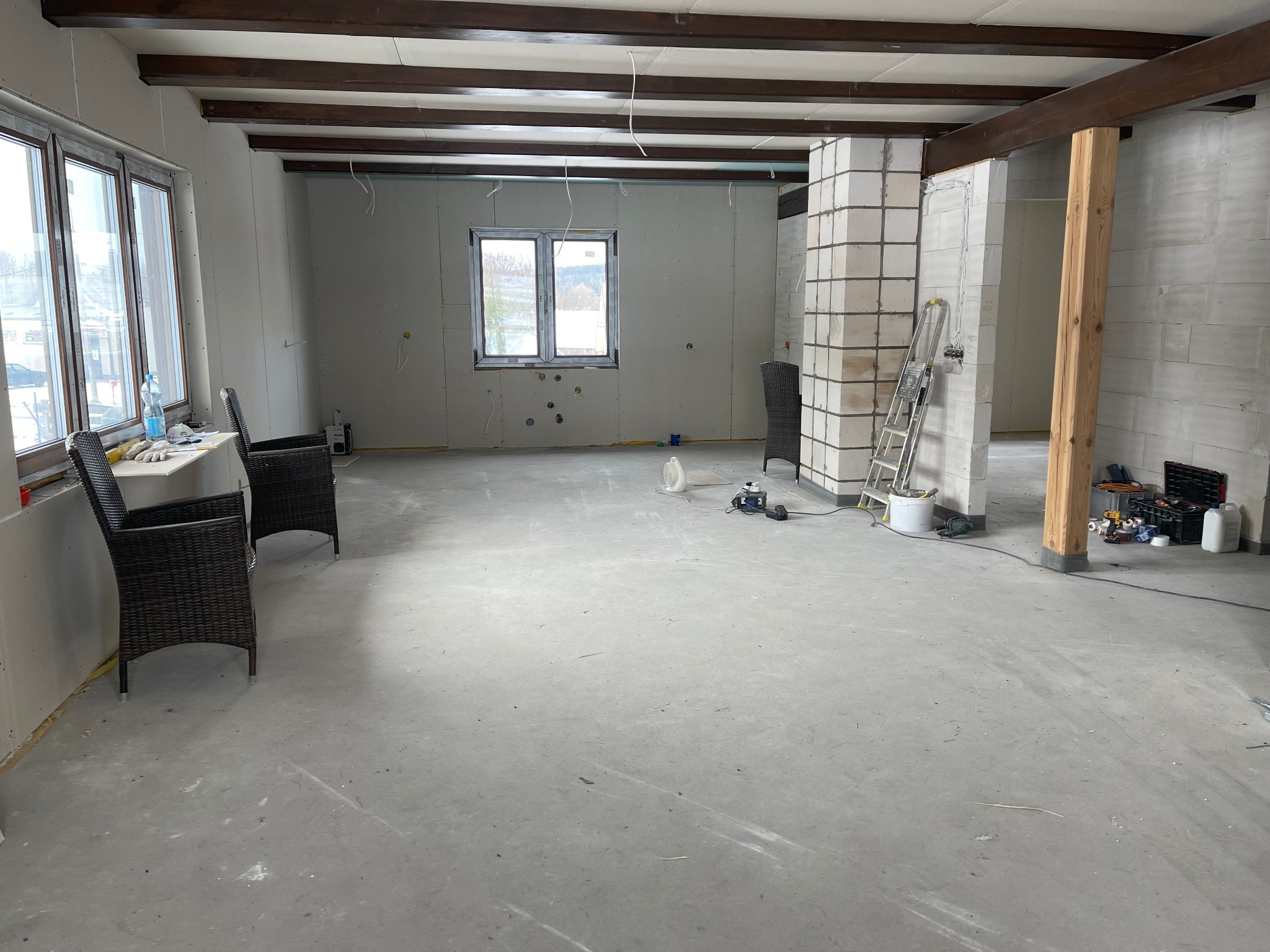 Wnętrze w trakcie prac wykończeniowych: ściany z płyt kartonowo-gipsowych, surowe betonowe podłoże, widoczne instalacje elektryczne, drewniane belki stropowe, okna z widokiem na zewnątrz, krzesła...